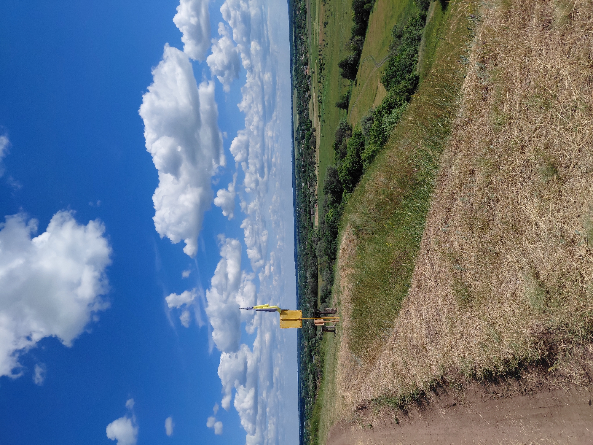 A dirt road curves along the left side of the frame, leading uphill. The foreground is dominated by dry, brown grass and some green tufts. A bright yellow signpost stands on the grassy slope, topped with a Ukrainian flag. Behind the signpost, a wooden structure resembling a bench or table with log supports is visible. The middle ground features a gently sloping green landscape dotted with trees and a small village. The background shows a vast, flat expanse under a bright blue sky with scattered white, puffy clouds. The scene is set in Ustyvytsia, Ukraine.