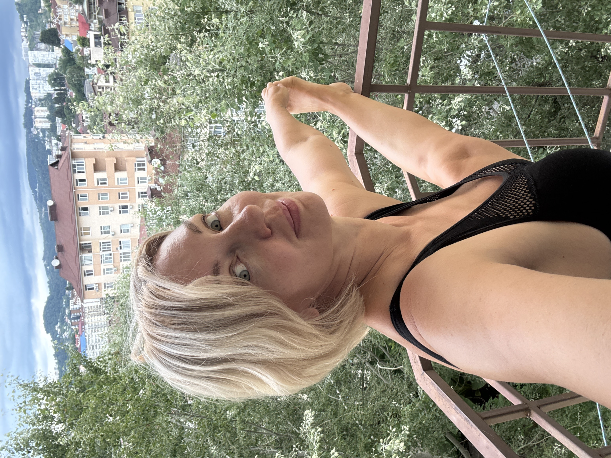 A woman with blonde hair and green eyes is on a balcony, holding her leg up over the railing. She is wearing a black top. In the background, there are buildings and trees, with mountains in the distance, suggesting a city location like Sochi, Russia. The sky is overcast.