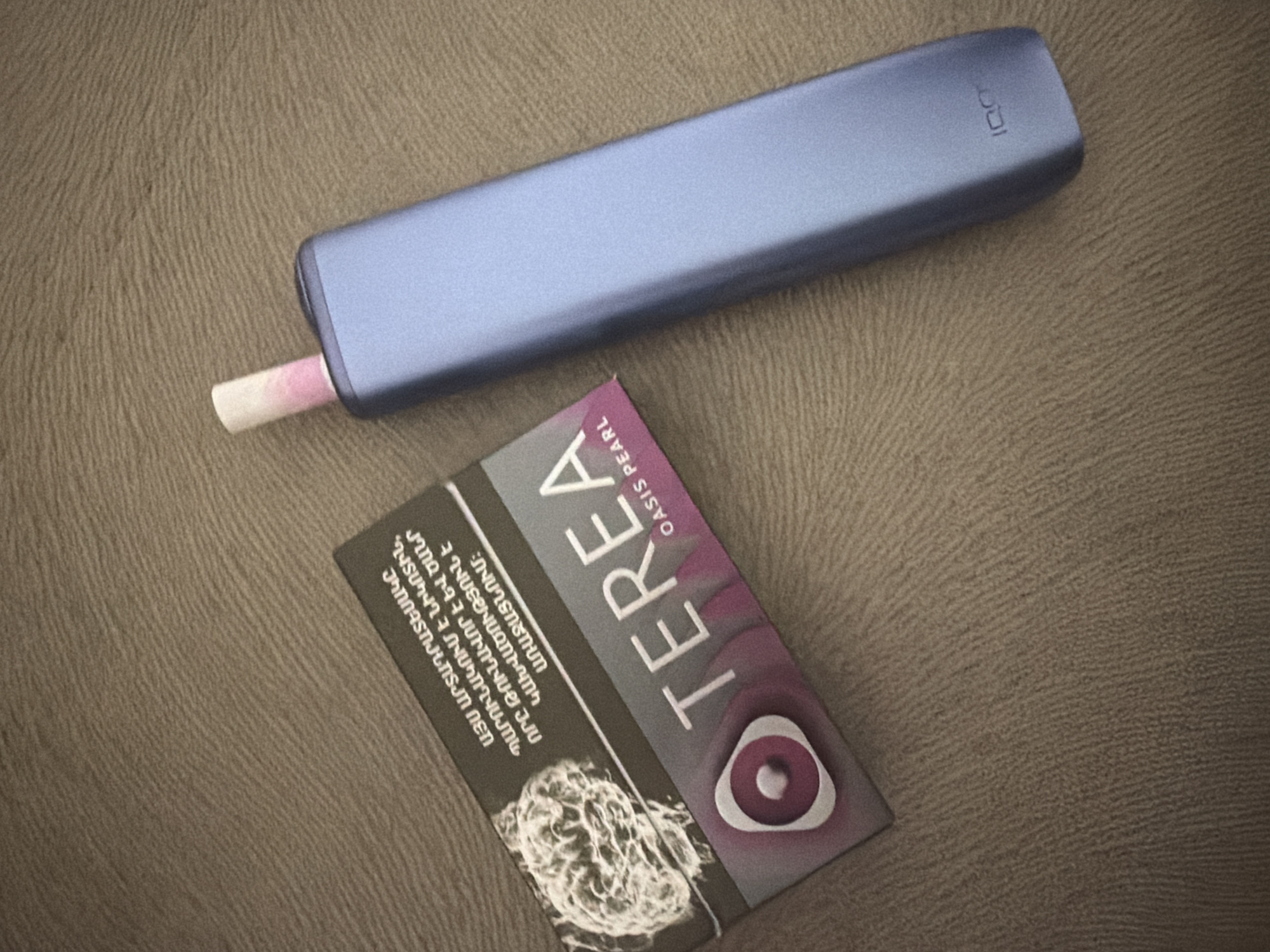 The image shows a blue IQOS device with a used TEREA Oasis Pearl stick inserted, alongside a pack of TEREA Oasis Pearl heatsticks, all resting on a textured brown surface. The TEREA packaging includes Armenian text and a design featuring a circular element. The image does not allow for the determination of location, as no visible elements indicate Yerevan, Armenia or any other specific place.