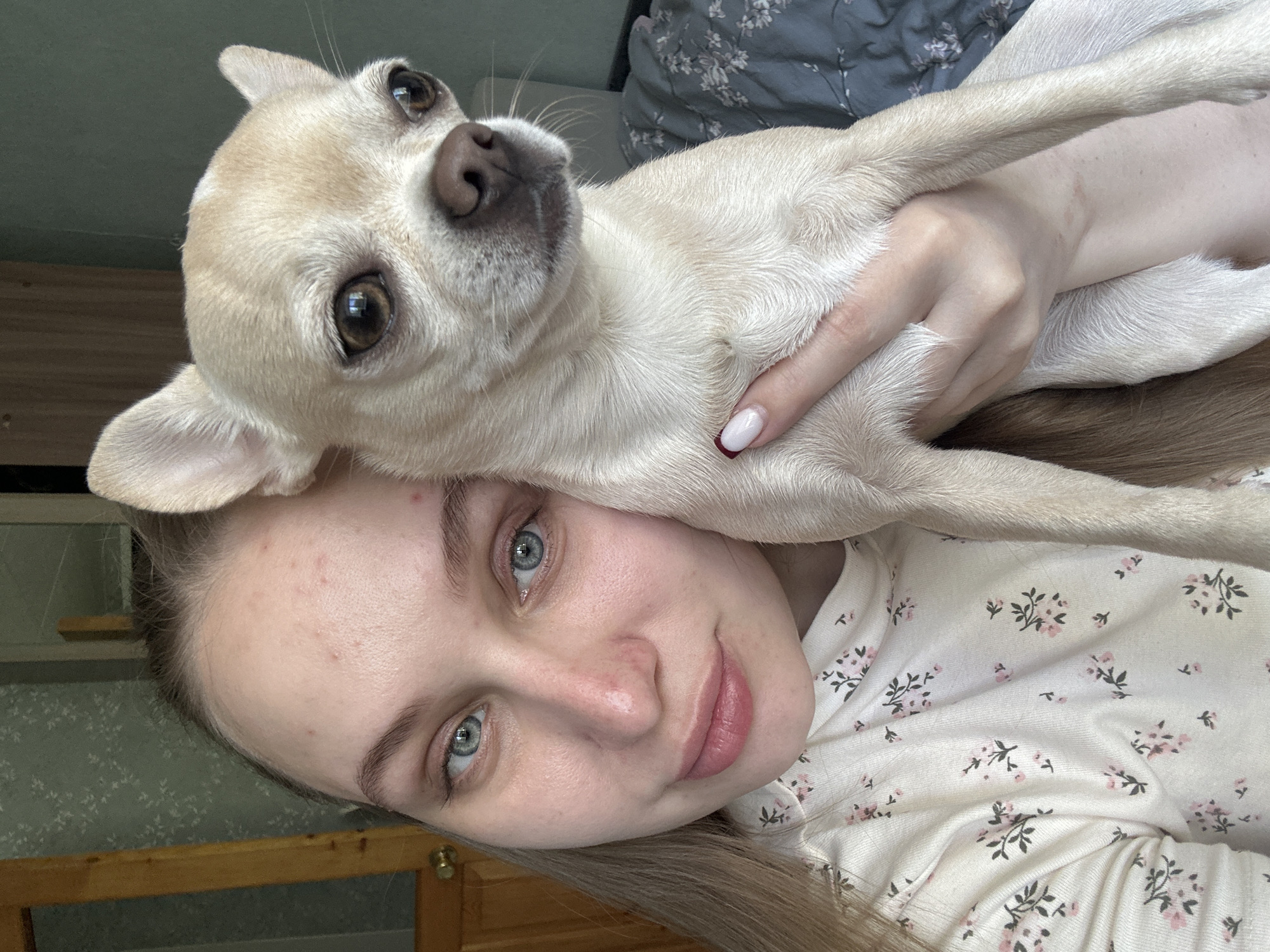 A light-skinned woman with blonde hair and blue eyes is holding a light beige chihuahua dog close to her face. The woman is wearing a white shirt with small pink floral patterns. She has her hand around the dog's chest. The photo appears to be taken indoors, possibly on a bed. The location is Chaikovsky, Russia.