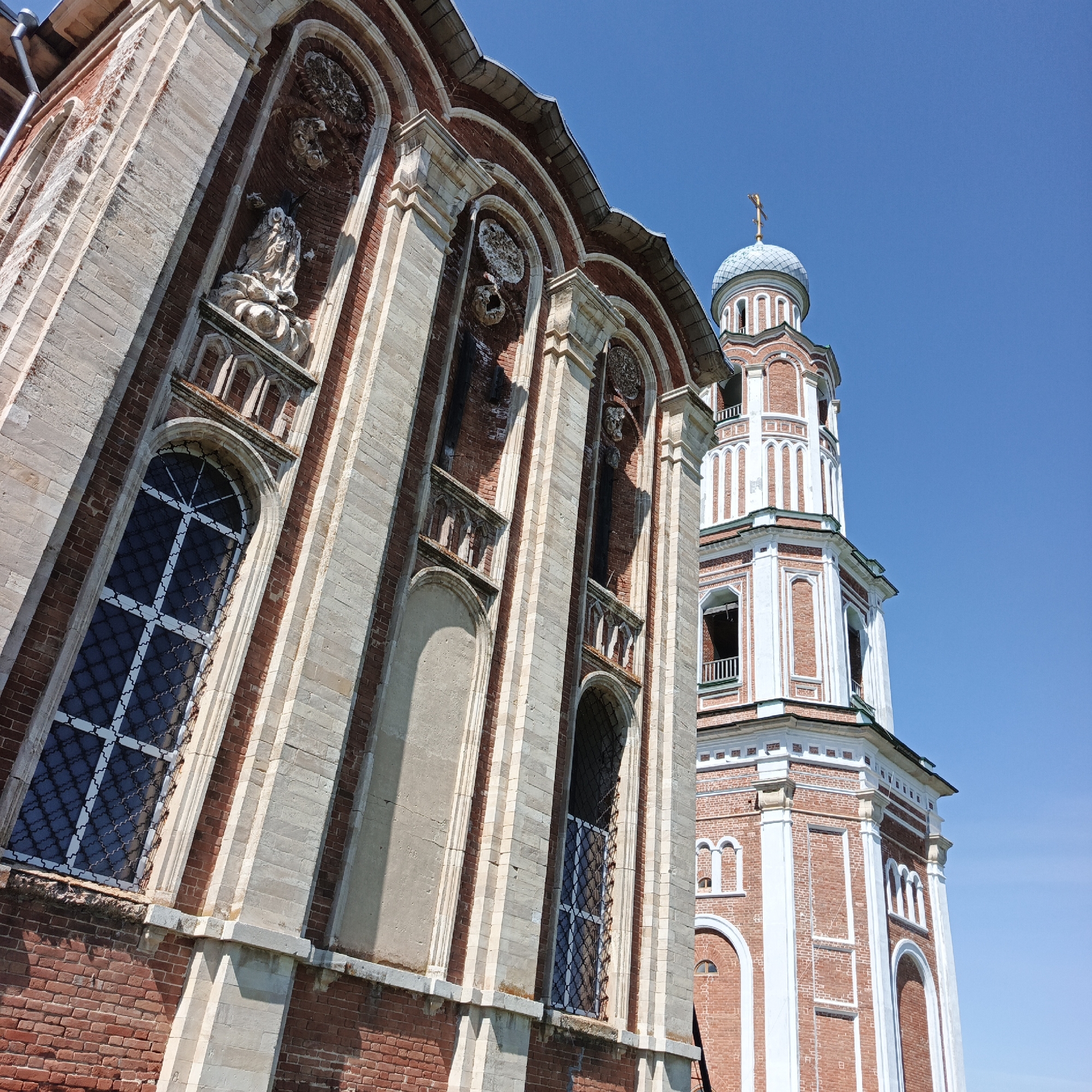 The image shows the architectural facade of a red brick church with white trim, featuring a tall bell tower topped with a blue dome and a golden cross. The church has arched windows with dark glass and ornate metal grates. Niches are visible on the facade, one containing a weathered statue. The architecture suggests a Russian Orthodox style. The scene is set against a clear blue sky. The location is identified as the city of Тугустемир, Russia. There are no people or animals present in the image. The lighting indicates a sunny day. The overall condition of the building appears to be well-maintained, though some weathering is visible on the stone elements and the statue.