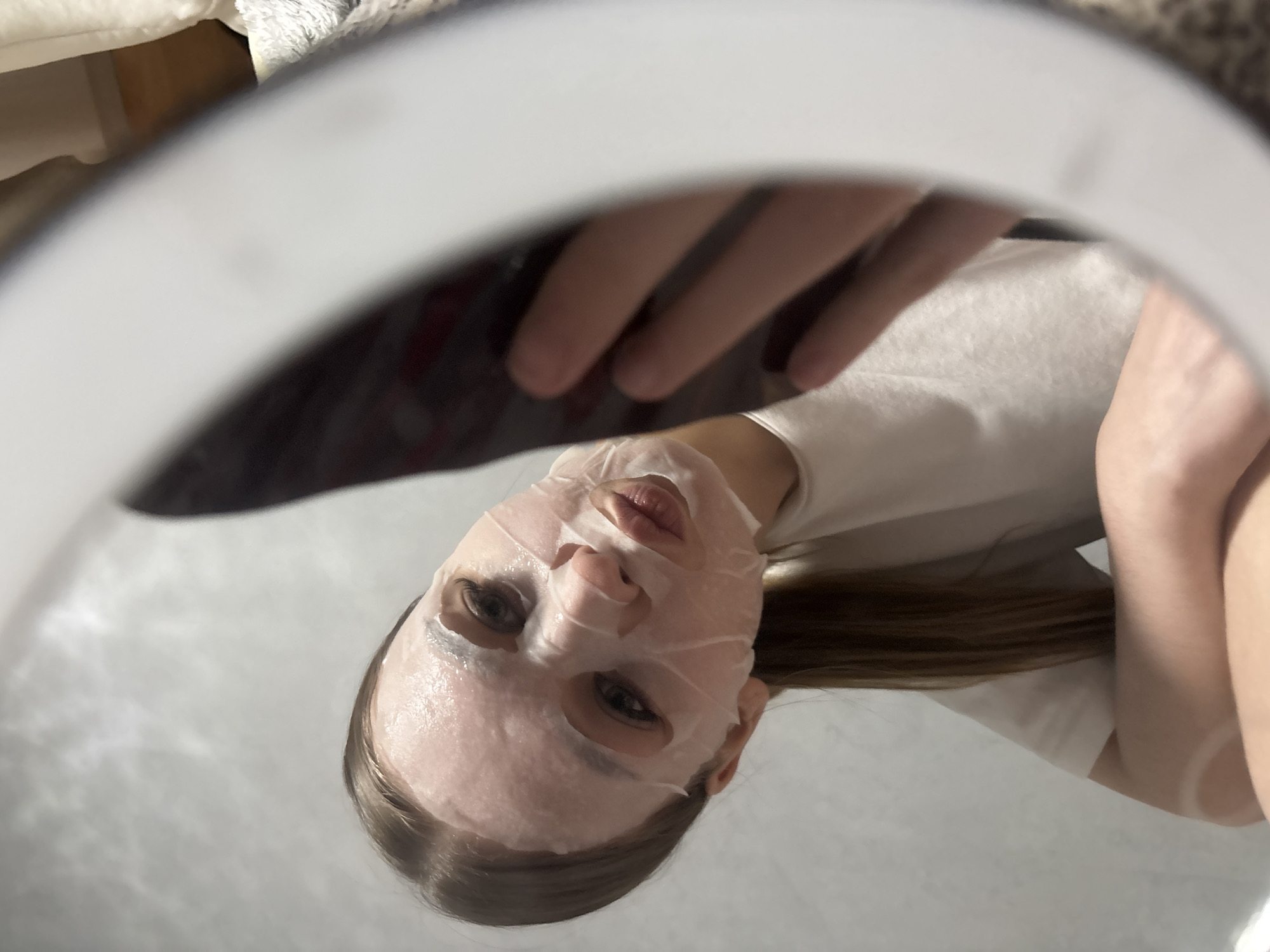 A woman is wearing a sheet face mask. The picture appears to be taken with the camera within a ring light. The woman is wearing a white t-shirt. The location is not evident. The city of Chaikovsky, Russia is not evident.