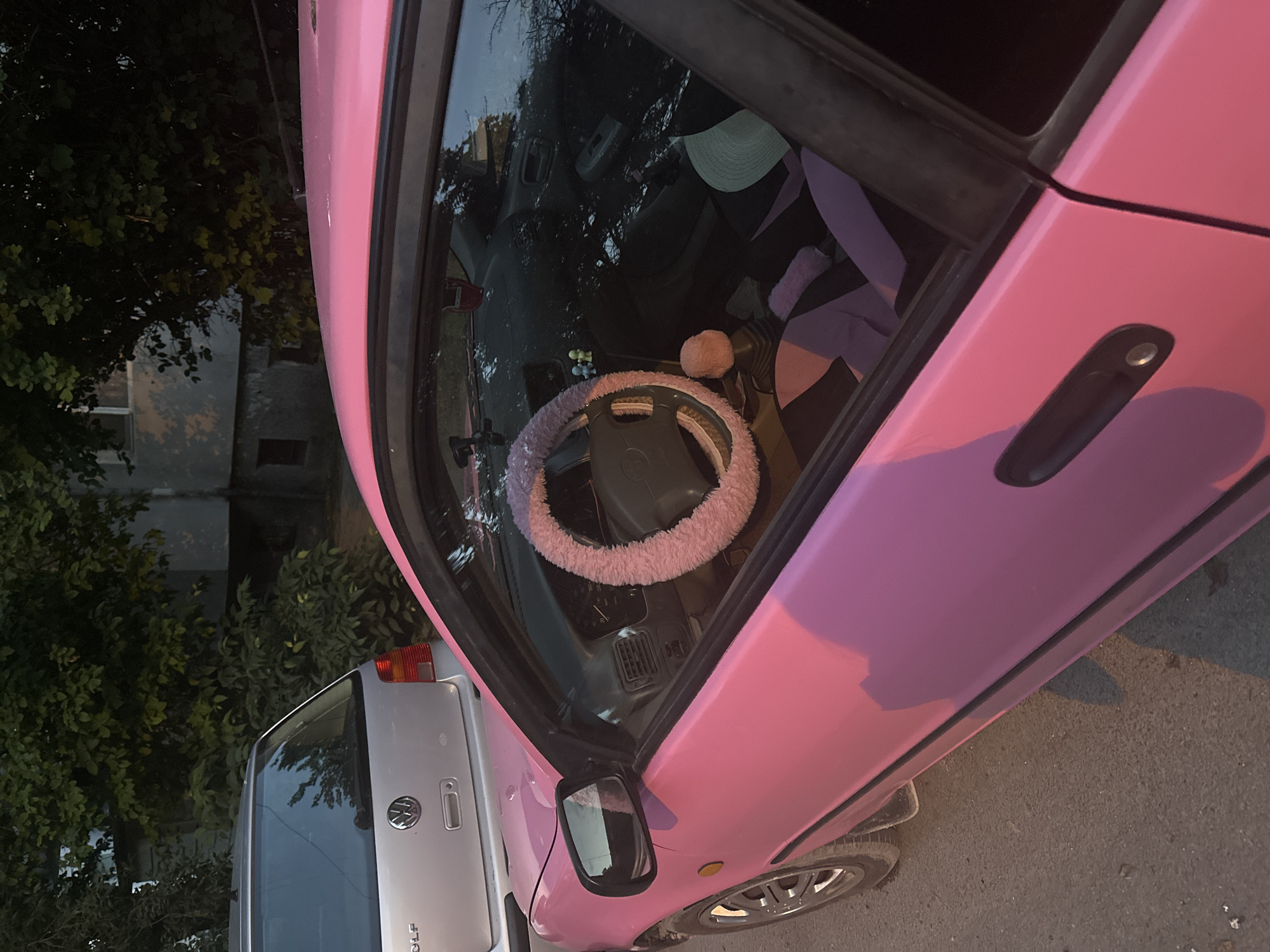 The image depicts a static street scene in Varna, Bulgaria, featuring two parked vehicles and a background environment. In the foreground, positioned to the right, is a bright pink automobile, specifically a Toyota (likely a Corolla based on the steering wheel emblem and dash design). The driver's side of the pink car is prominently visible, showcasing its matte pink exterior paint. The front left wheel, side mirror, and door handle are identifiable. Through the open driver's side window, the car's interior is detailed: a fluffy light pink cover adorns the steering wheel, a similar fluffy pink cover is on the gear shift knob, and the seats are fitted with black and light pink fabric covers. Small decorative figurines are present on the dashboard, and an air freshener hangs from the rearview mirror. A light-colored hat rests on the passenger seat.
Behind the pink car, and partially obscured, is a silver Volkswagen Golf, specifically identifiable as a Mk4 generation by its rear tailgate, taillights, and the "GOLF" inscription beneath the Volkswagen emblem.
The ground beneath the vehicles is a paved asphalt surface. In the background, behind the silver Golf, there is dense green foliage from trees and shrubs. Partially visible through the greenery is a multi-story building, appearing grey with subtle architectural details, suggesting a residential or commercial structure in an urban setting. The scene is illuminated by natural light, casting subtle shadows and highlighting the colors of the vehicles. No people or active interactions are visible in the frame.