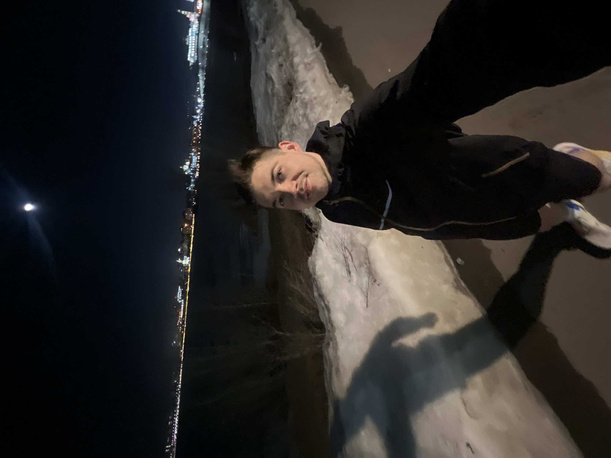 A sideways, medium-shot selfie of a fair-skinned male with light brown hair, wearing a dark jacket and white sneakers, taken at night in Kazan, Russia. The male is turned slightly toward the camera with a mild smile. His left shoulder and part of his right side are visible. He is standing on a concrete surface with patches of snow and bare branches nearby. His shadow is cast on the ground. In the background, across a body of water, is a cityscape illuminated with numerous lights. A bright, small circle, possibly the moon, is visible in the dark sky to the left. The image appears to be shot from a slightly elevated position.