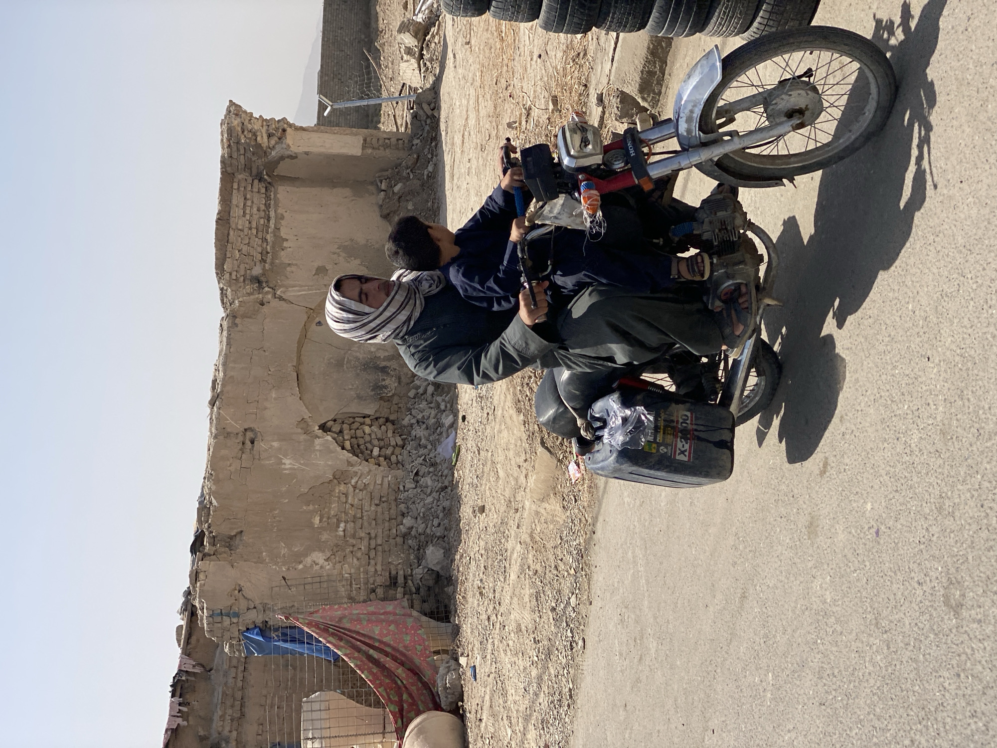 In Kandahar, Afghanistan, a person wearing a headscarf and traditional clothing rides a motorcycle with a child seated in front of them. The motorcycle is carrying a black fuel container labeled "X-2000." In the background, partially ruined brick buildings and debris are visible under a hazy sky. The scene appears to be set on a dirt road or pathway.