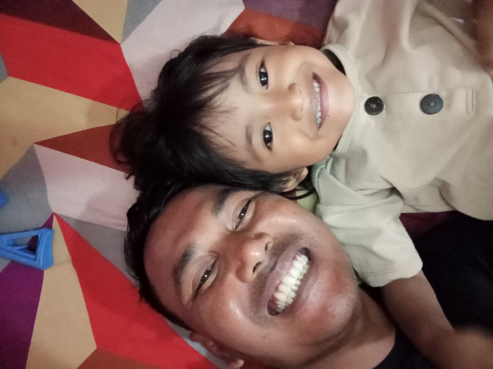 In Bloro, Indonesia, a selfie captures a man and a young child lying down, possibly on a bed with a geometric patterned spread. The man, with dark hair and a fair complexion, is in the foreground, his face angled towards the camera with a wide, toothy smile. He is wearing a dark shirt. The child, with dark hair and a round face, is positioned above and behind the man, also smiling and looking towards the camera. The child is wearing a light-colored shirt with two dark buttons. The background features a colorful, geometric pattern. A blue plastic object, possibly a toy, is visible on the left side of the frame. The lighting is soft, suggesting an indoor setting.