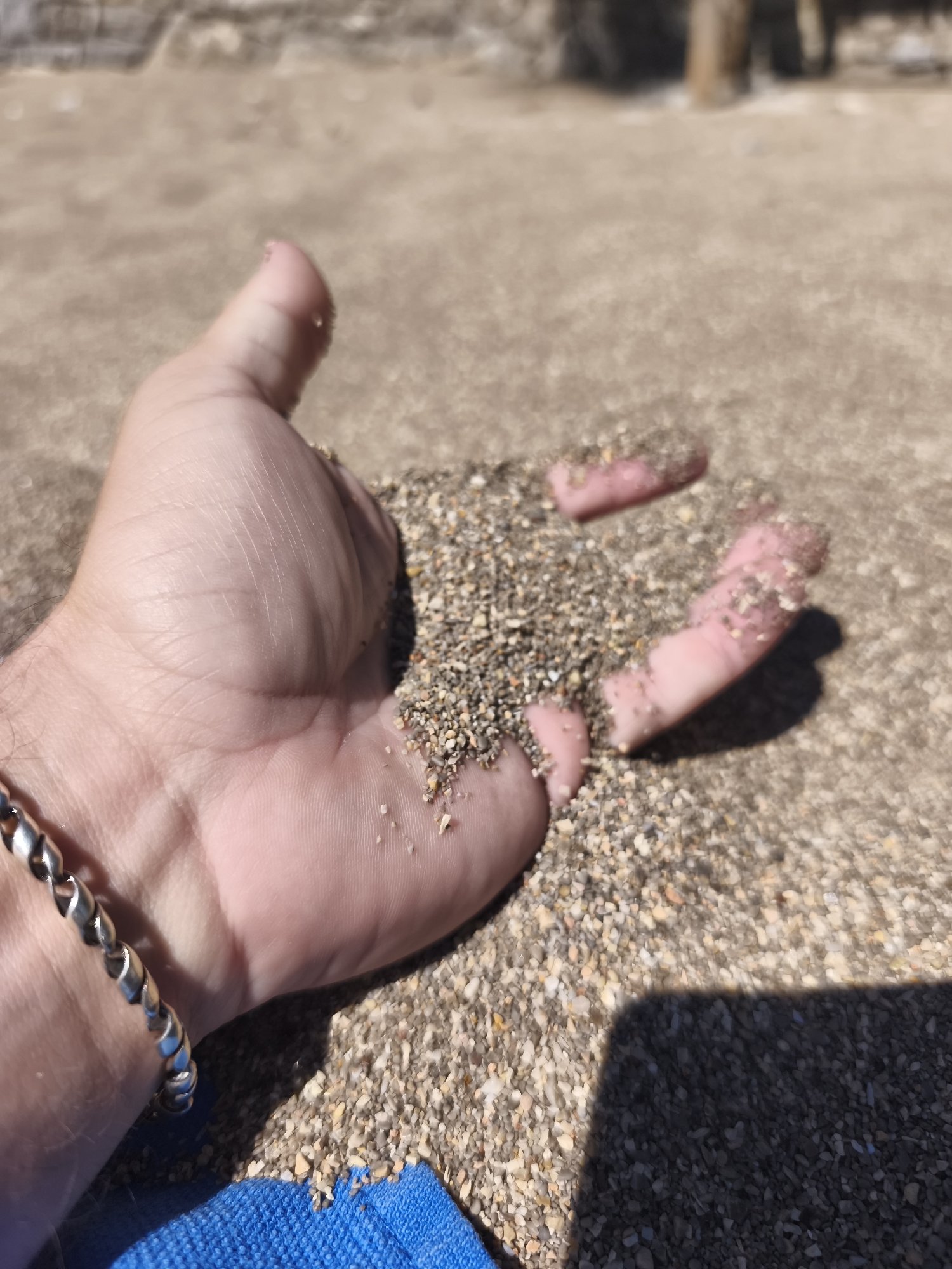 The image shows a person's hand filled with sand. The hand is holding the sand against a backdrop of more sand. The person is wearing a silver bracelet. A small piece of blue fabric is visible at the bottom left corner of the image. The location is Budva, Montenegro, likely on a beach. The sand appears to be a mix of light and dark grains. There is a wooden pole visible in the blurred background. The lighting suggests it is a sunny day.