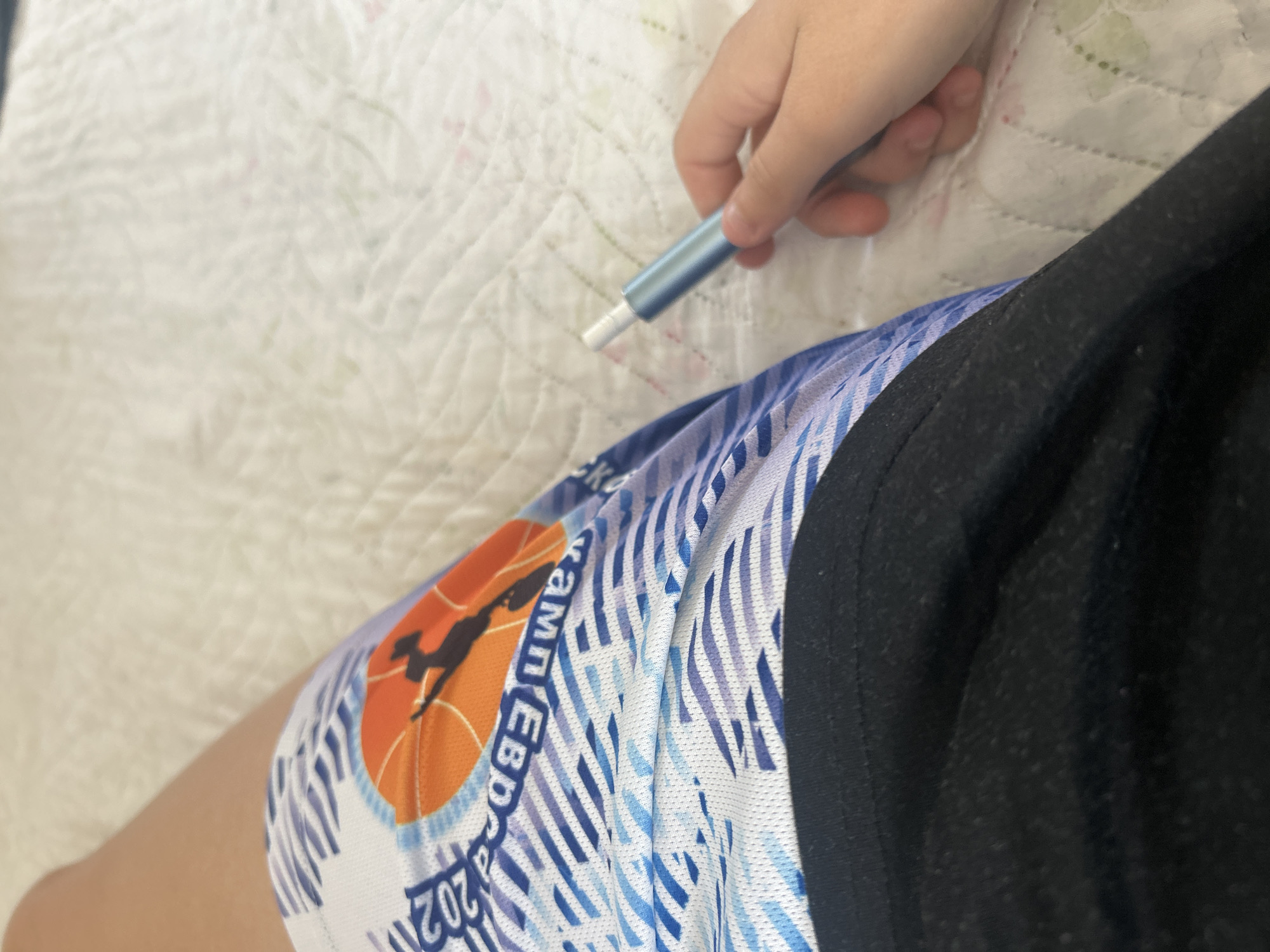 The image shows a person's leg wearing a blue and white patterned garment with the text "KAM EBD 2023". A hand is holding a light blue cylindrical object. The background is a white quilted surface. There is no indication of the city of Shumen, Bulgaria in the image.