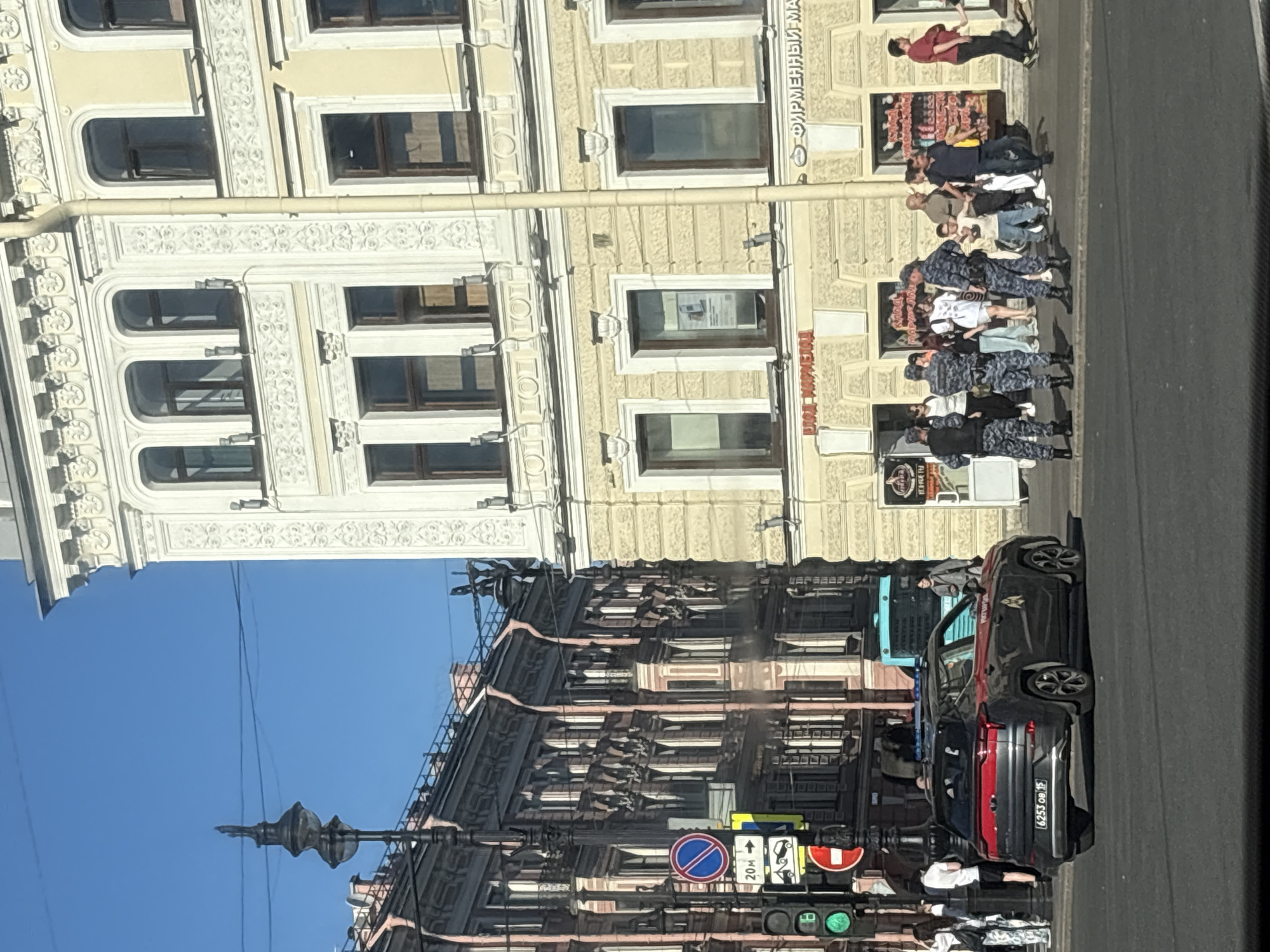 This daytime image depicts a busy urban street scene on Nevsky Prospekt, specifically near number 30, in Saint Petersburg, Russia, under a clear blue sky.
In the foreground, a dark gray SUV, identified as a Rosgvardiya (Russian National Guard) vehicle by its side markings and blue flashing lights, is parked on the asphalt street, facing left. The vehicle's rear license plate, "6253 OB 15," is clearly visible. Five uniformed Rosgvardiya officers, dressed in dark blue camouflage uniforms with black tactical vests and hats, are standing on the sidewalk to the right of the police vehicle. They are positioned in a loose group, observing the surroundings.
A diverse group of civilians, casually dressed in summer attire, are present on the sidewalk and near the street. Some are walking, while others are standing near the storefronts. Notable civilians include a woman with a patterned shirt and dark pants walking left, a man in a red t-shirt walking right, and a child in light clothing within a group near the officers. A light blue bus is partially visible in the background, further down the street behind the Rosgvardiya vehicle.
The right side of the frame features a multi-story building with a light yellow/cream and white facade, characterized by ornate stucco decorations, arched windows on upper floors, and rectangular windows on lower floors. The ground floor hosts storefronts, with visible Russian signage including "ВЛАД МАРМЕЛАД" (Vlad Marmelad) and "ФИРМЕННЫЙ МАГАЗИН" (Company Store). A light-colored drainpipe runs vertically down the building's facade.
On the left side, further back, another multi-story building with a darker, reddish-brown facade and numerous windows is visible, also featuring ornate architectural details. A tall, black, ornate lamppost with a small figure or statue on its top stands prominently in the mid-ground on the left. Traffic lights showing a green signal are visible below the lamppost, alongside various street signs: a blue circular "No Stopping/No Parking" sign, a blue circular sign with a white arrow indicating "20м" to the right, and a yellow "Tow Away Zone" sign.