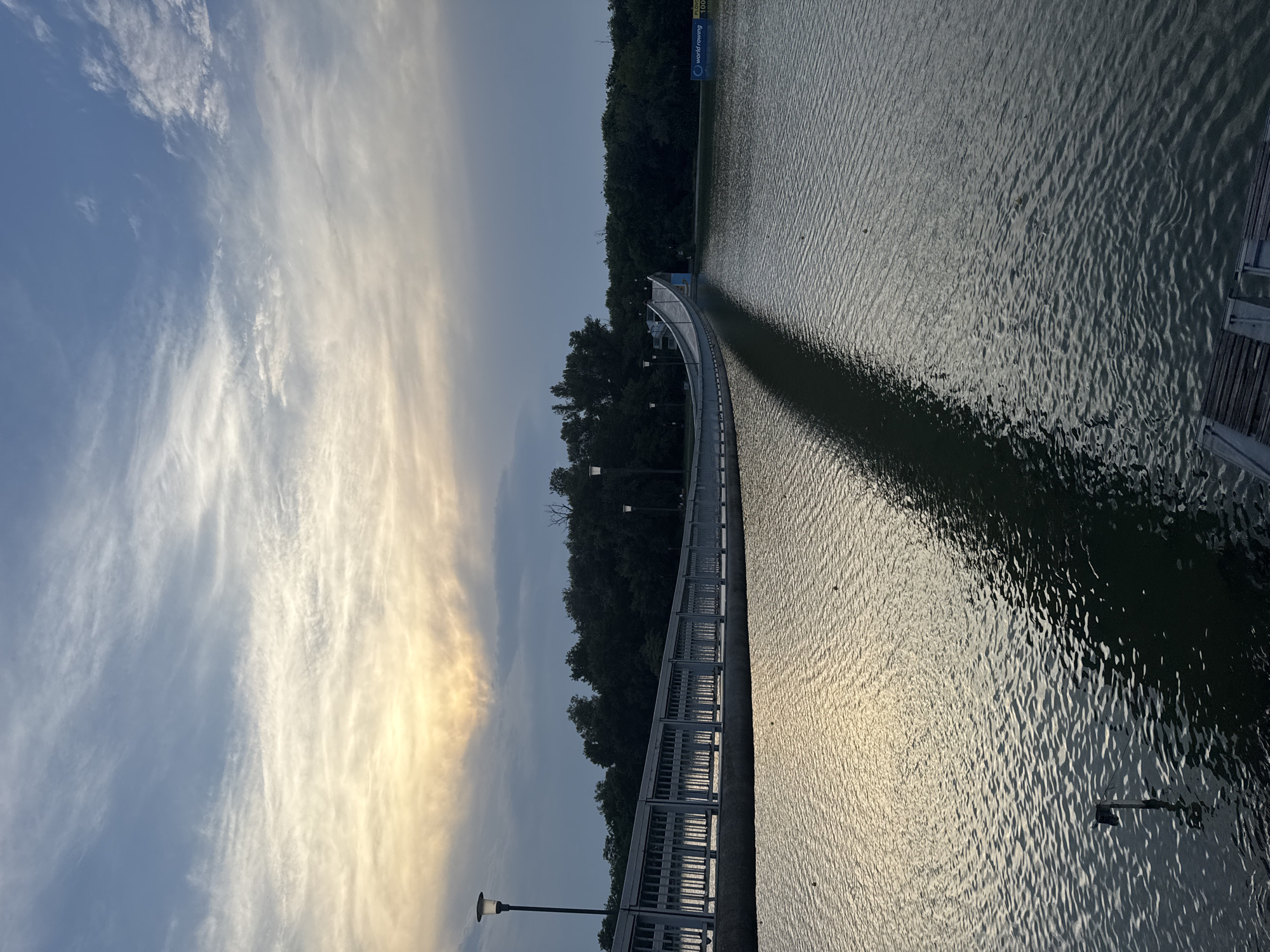 The image depicts a long, arched pedestrian bridge extending over a body of water, likely a lake or river. The bridge has a light-colored railing and is illuminated by lampposts spaced at intervals. A dense line of trees borders the opposite bank of the water. The sky above is partly cloudy with a soft glow of sunset or sunrise, casting a reflection on the water's surface. A shadow of the bridge stretches across the water. In the background, a blue sign with white lettering, possibly advertising a rowing event, is visible on the right side. The scene is calm and appears to be set in an outdoor recreational area. This location is identified as the Rowing Canal in Plovdiv, Bulgaria.
