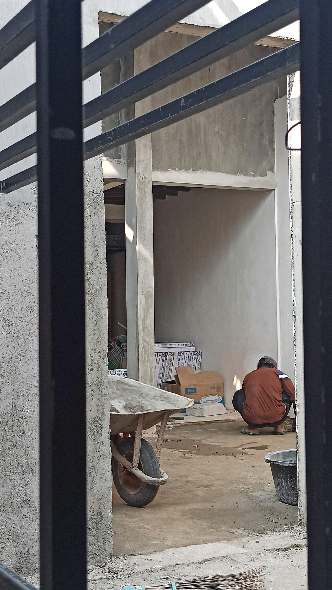 The image depicts an outdoor construction site in Ngumpul, Indonesia. In the foreground, a black metal fence partially obstructs the view. To the left, a rough concrete wall is visible. A wheelbarrow with a single wheel, its metal basin containing some materials, is positioned in the mid-left of the frame. The ground is a concrete surface, appearing somewhat damp in places, with scattered debris. In the background, a person wearing a brown long-sleeved shirt and dark pants is crouched down, facing away from the camera. They appear to be engaged in some activity on the ground. To their right, a black plastic tub is situated. Behind the person, stacks of what appear to be construction materials, possibly tiles or boards, are leaning against a wall, along with cardboard boxes. The architectural elements suggest an unfinished building, with exposed concrete walls and structural beams. Sunlight casts shadows, indicating it is daytime.