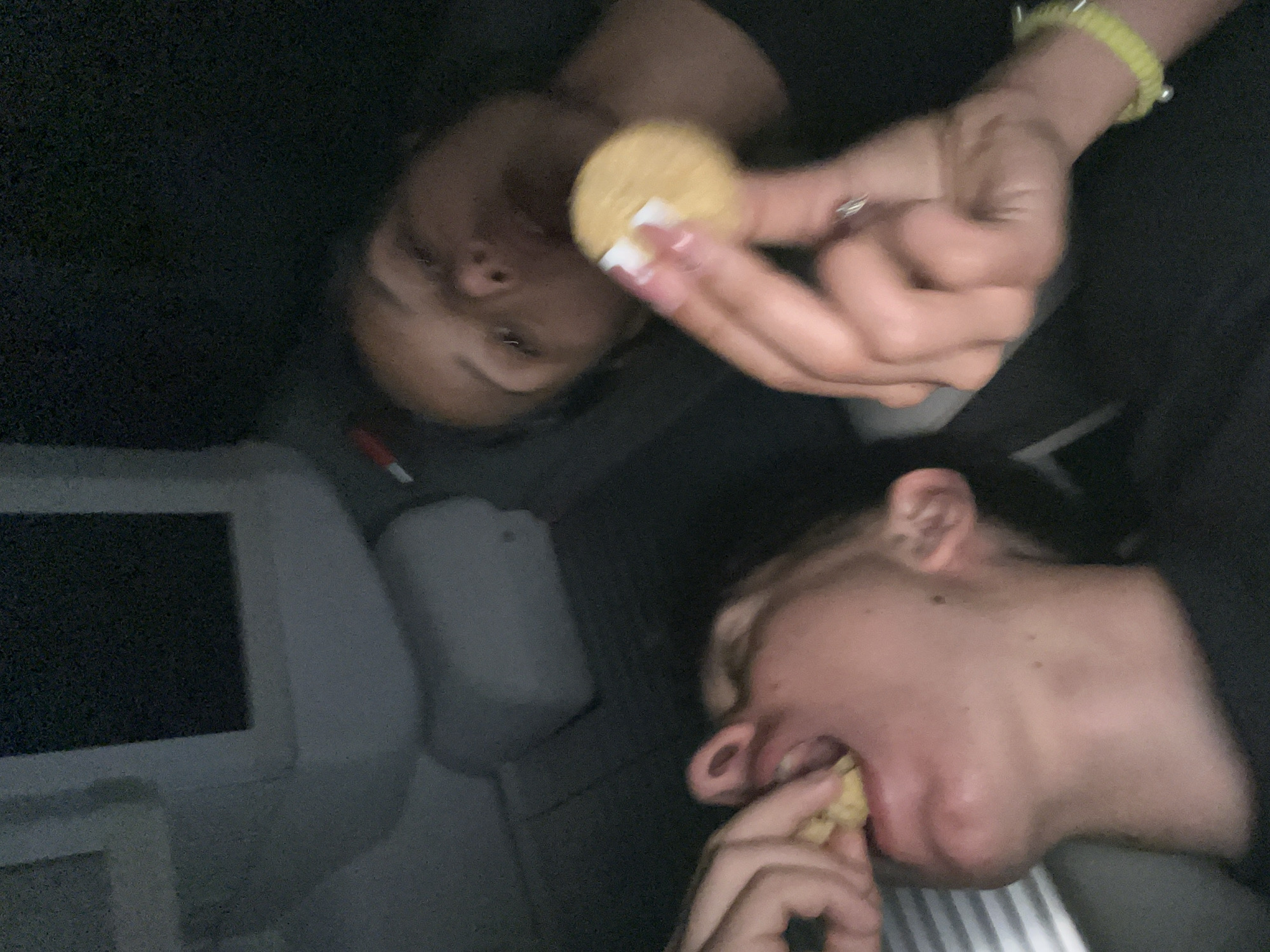 Two people are visible in the dark indoor setting. One person with long fingernails, some painted white, is holding a round, yellow cookie. The person below is eating a similar cookie. A yellow bracelet is on the wrist of the person holding the cookie. An airplane entertainment screen and what appears to be a pillow are visible in the lower left corner of the frame. No information indicates a connection to Popovitsa, Bulgaria.
