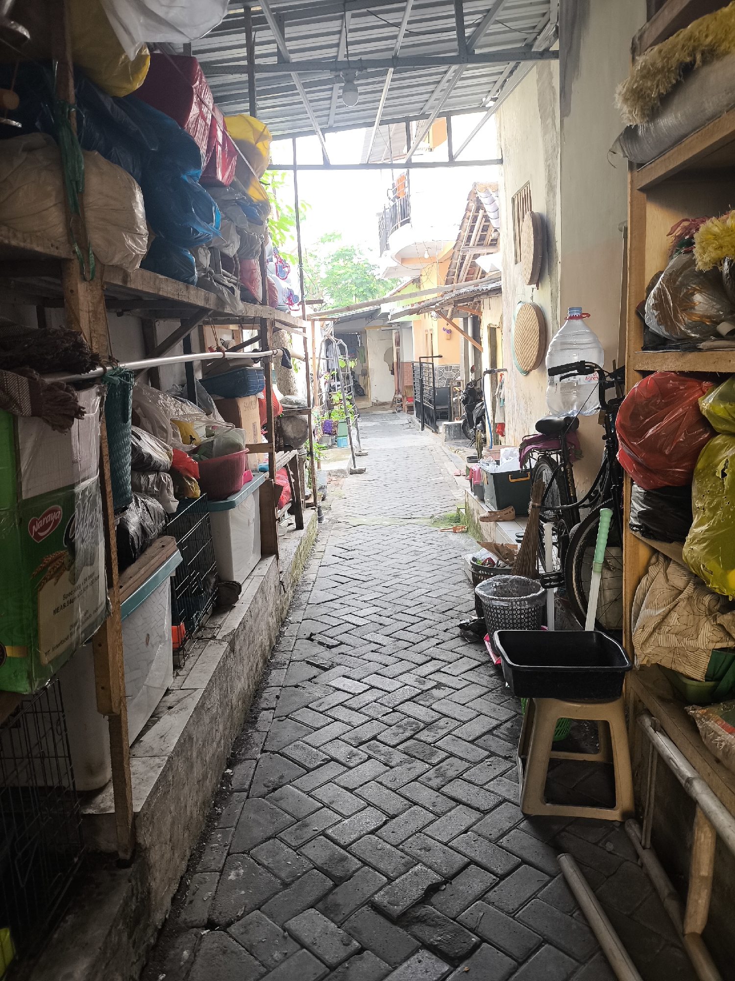 The image depicts a narrow, cobblestone alleyway in the Citrodiwangsan area of Indonesia. The alley is lined with shelves overflowing with various items, including large sacks of goods, plastic containers, and what appear to be cleaning supplies. On the left side, a shelf holds a green box labeled "Naraya" and several other assorted items in sacks and plastic bags. The right side of the alley features more shelving with items, including red and yellow plastic bags. A bicycle with a large water jug on its back is parked against the wall. A broom and a dustpan are also visible near the bicycle. The alley has a covered roof structure, suggesting it might be a walkway or a storage area within a residential or commercial complex. The ground is paved with interlocking bricks and shows signs of moisture. The overall scene conveys a sense of cluttered storage and functional utility within a tightly packed urban environment.