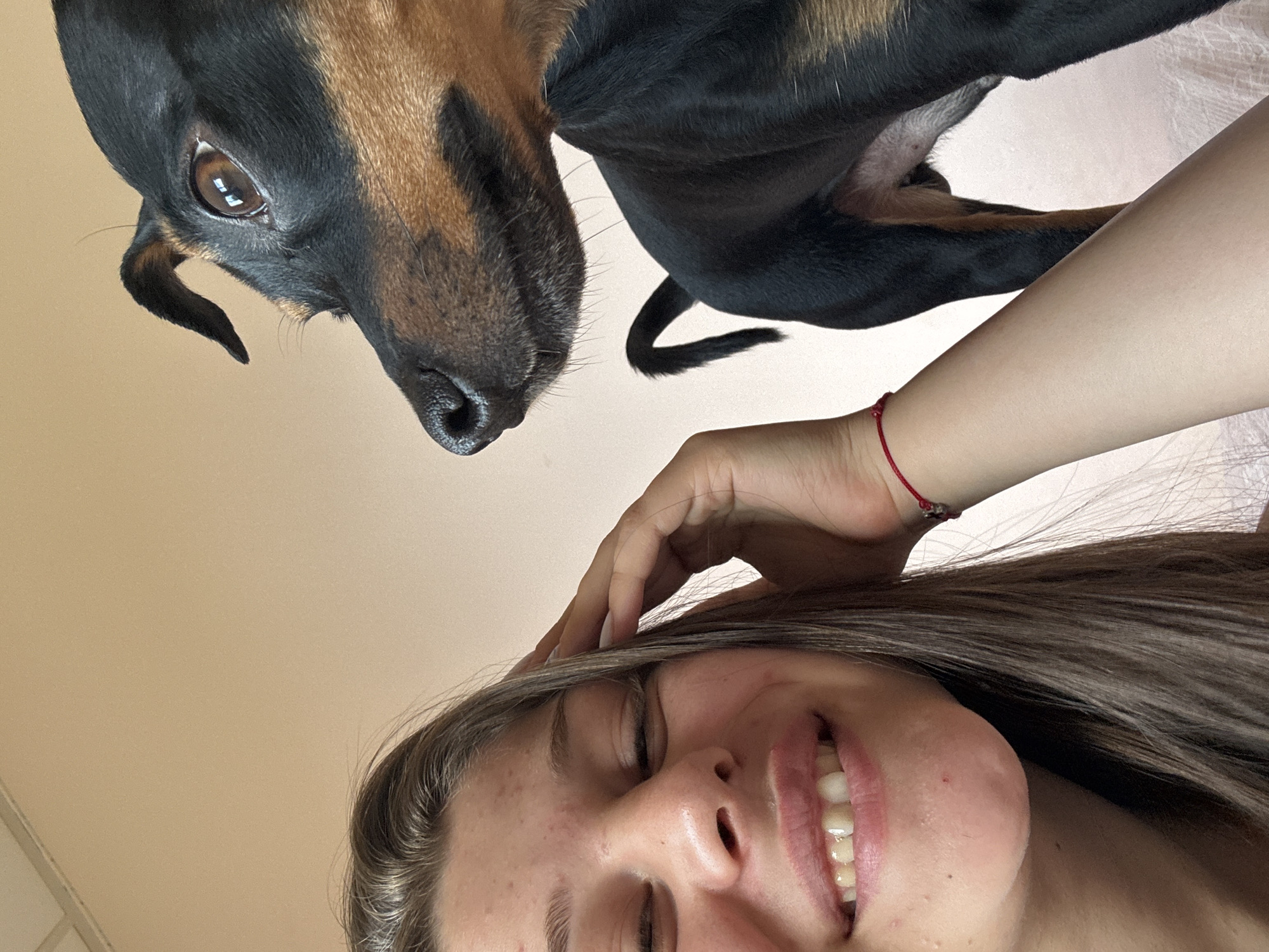 The media file is a close-up, low-angle selfie featuring a young woman and a dog. The woman is positioned in the foreground, with her head tilted back and eyes closed, smiling broadly. Her long, dark hair frames her face. Her hand is resting on her head, with a thin red string bracelet visible on her wrist.
In the background, a dog's head and upper body are visible, also angled down towards the camera. The dog has dark fur with tan markings and its eye is clearly visible, reflecting light. Its nose is pointed towards the woman. The background appears to be a plain, light-colored wall. The lighting suggests it is daytime. The overall mood is playful and intimate, capturing a moment between a person and their pet. There is no visible text in the image.