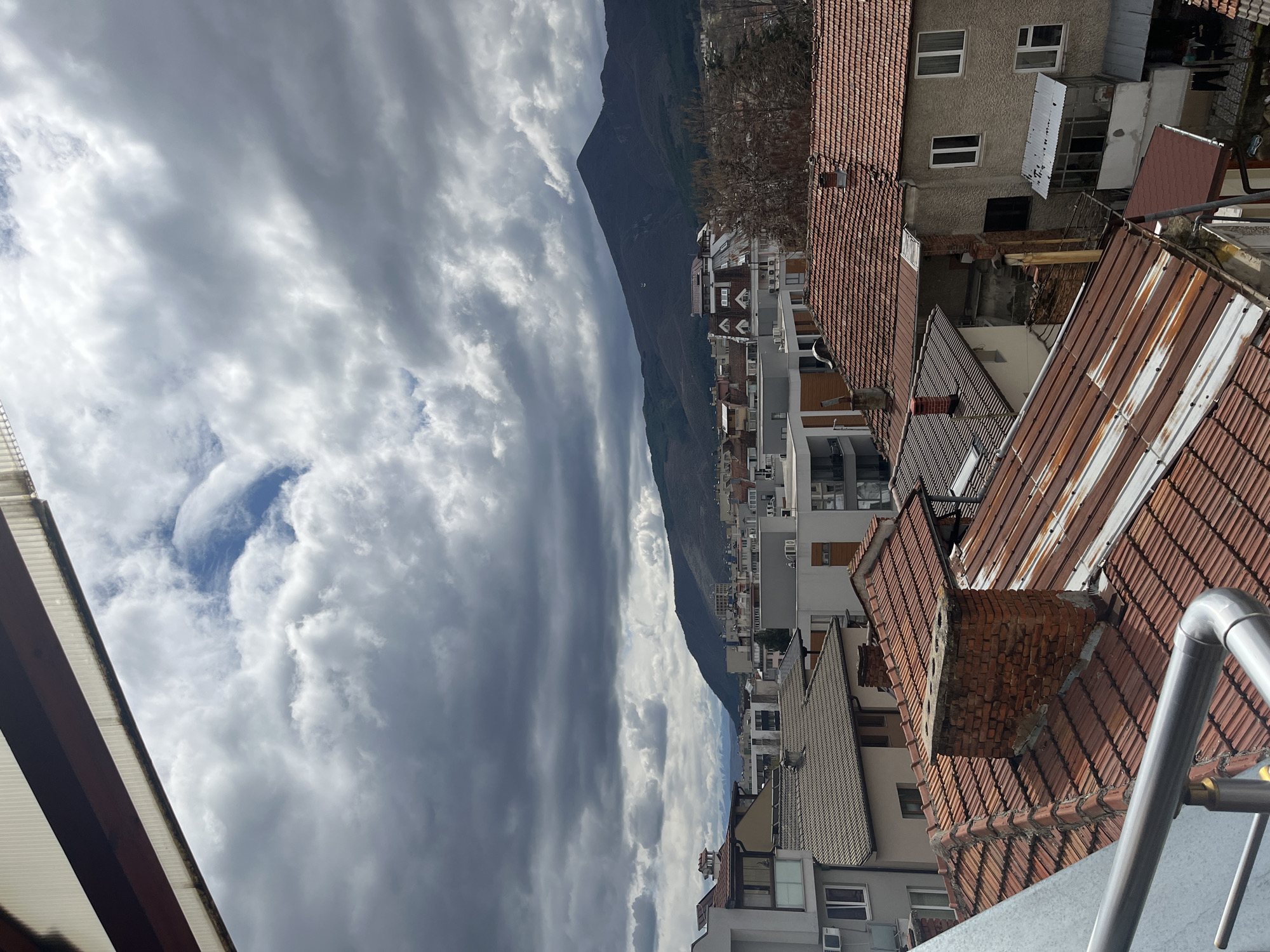 The image shows a cityscape in Goce Delchev, Bulgaria, viewed from an elevated position, likely a balcony or upper floor. The foreground features residential buildings with tiled roofs of varying ages and conditions, including some with brick chimneys. Further in the background, there are multi-story apartment buildings and commercial structures. A mountain rises prominently in the distance, separating the urban area from the sky. The sky is filled with heavy, layered clouds. The image is rotated, with the sky appearing on the left and the urban landscape on the right. A metal railing is visible in the bottom-right corner.