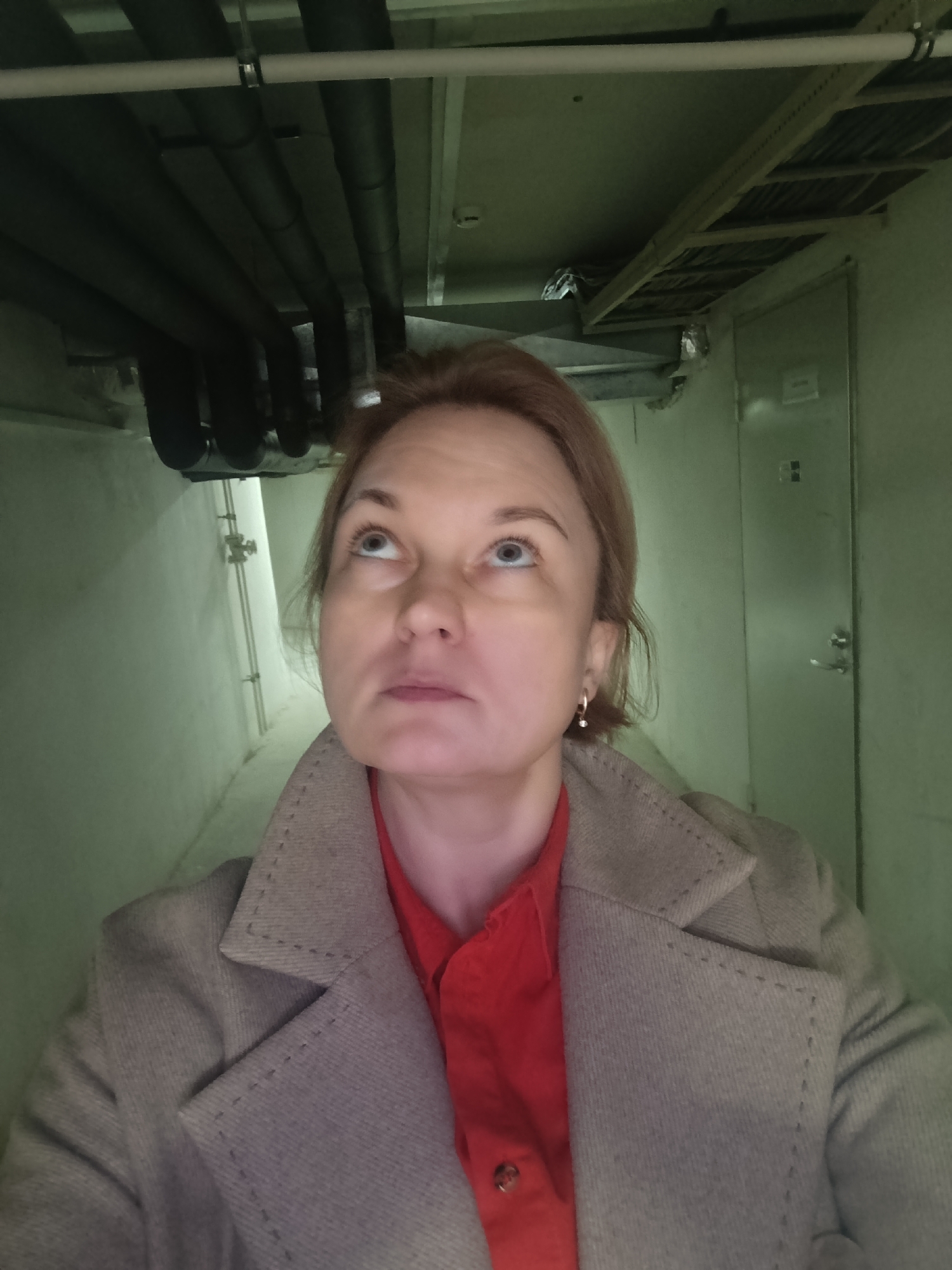 A woman with light brown hair and blue eyes is captured in a head-and-shoulders portrait, looking upwards with a curious or contemplative expression. She is wearing a vibrant red collared shirt beneath a light grey or beige coat featuring distinct, raised stitching on the lapels and pockets. A small, simple hoop earring is visible on her right ear.
The setting appears to be an indoor utility corridor or maintenance area, possibly a basement or a back-of-house space in Марковский, Russia. The walls are a muted green or grey color, and the lighting is somewhat dim and utilitarian. Above the woman, the ceiling reveals exposed infrastructure, including multiple dark, insulated pipes running horizontally, a lighter-colored pipe, and a metal cable tray or racking system. To the left, a white pipe with fittings runs vertically along the wall. To the right, a plain, light-colored metal door with a handle and what appears to be a small, unreadable sign is visible. The overall environment suggests a functional, non-public space. No clearly legible text is visible in the image.