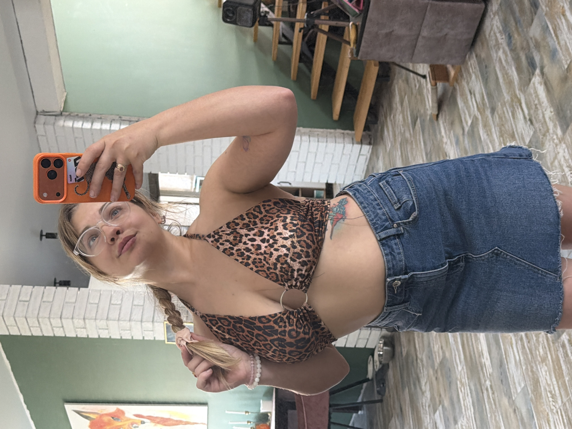 A woman is taking a selfie in a room. She is wearing glasses, a leopard print top, and a denim skirt. The room has white brick walls and a green ceiling. There are stairs and furniture in the background. There is no way to know if this photo was taken in Большая Шильна, Russia.