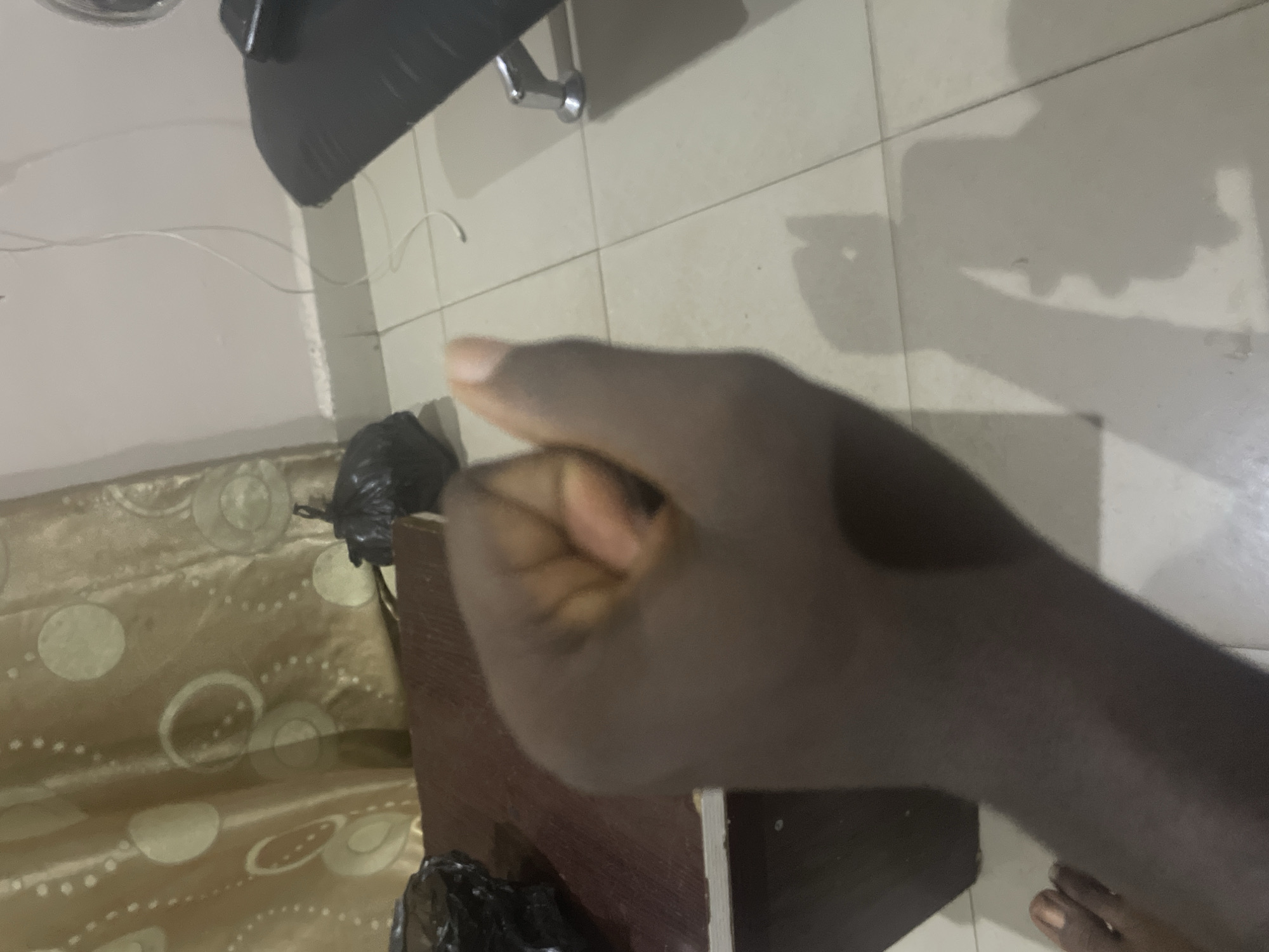 A first-person perspective shot of a person's right hand in a closed fist, with the thumb partially extended upwards. The hand is dark-skinned. In the background, a brown wooden surface is visible, with black plastic bags on top and to the left. Further in the background, a patterned curtain hangs, and a white electrical cord snakes down a light-colored wall. The floor is tiled in a pale color, and a shadow of the person's hand and arm is cast on it. A black chair with a metal leg is partially visible on the right. The scene is indoors, likely in a residential setting in Jalingo, Nigeria, given the context.