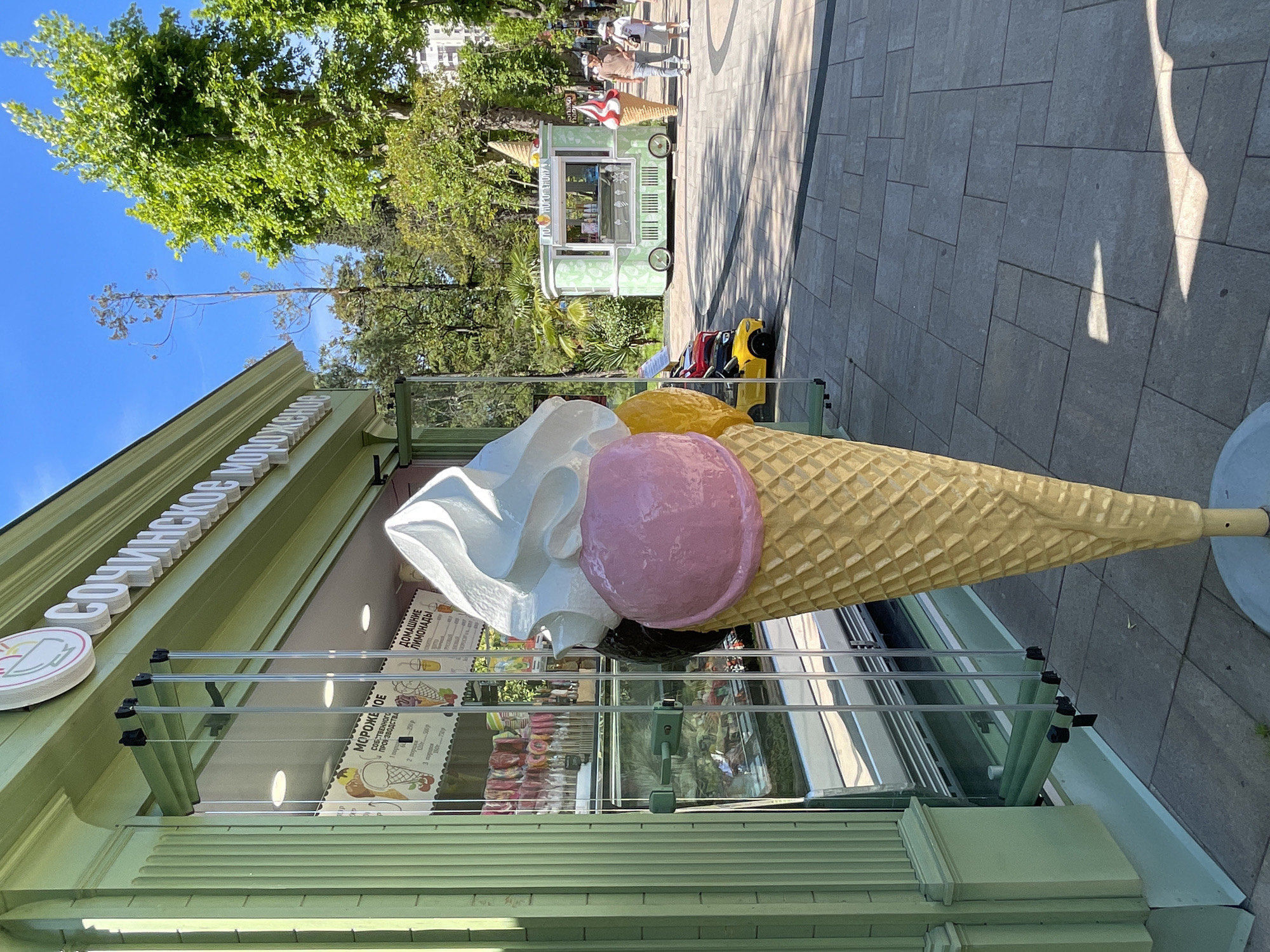 The image, taken in Sochi, Russia, captures an outdoor scene dominated by a large, colorful decorative ice cream cone. The cone, made of a waffle texture, holds three oversized scoops: a large white scoop resembling whipped cream or vanilla, a pink scoop, and a yellow scoop. It stands in front of a light green ice cream parlor or kiosk.
The parlor's facade features "СОЧИНСКОЕ МОРОЖЕНОЕ" (Sochi Ice Cream) in prominent white lettering, alongside a circular logo depicting an ice cream cone. Behind a clear protective screen, a menu board lists items such as "МОРОЖЕНОЕ СОБСТВЕННОГО ПРОИЗВОДСТВА" (Own Production Ice Cream) with prices for scoops (e.g., 1 scoop - 150₽, 2 scoops - 250₽, 3 scoops - 300₽) and by weight (e.g., 100g - 250₽). Various flavors are partially visible, including chocolate, vanilla, strawberry, pistachio, and caramel. "ДОМАШНИЕ ЛИМОНАДЫ" (Homemade Lemonades) are also advertised with their prices.
The scene takes place on a bright, sunny day, indicated by the clear blue sky and strong shadows cast on the light gray paved pathway. Lush green trees line the pathway, suggesting a park or pedestrian area. In the background, several people are seen walking, and a smaller, mobile ice cream cart, also green and displaying "МОРОЖЕНОЕ" (Ice Cream), is parked. Near this cart are a few brightly colored ride-on toy vehicles, including a yellow car. The overall impression is one of a pleasant, leisurely day.