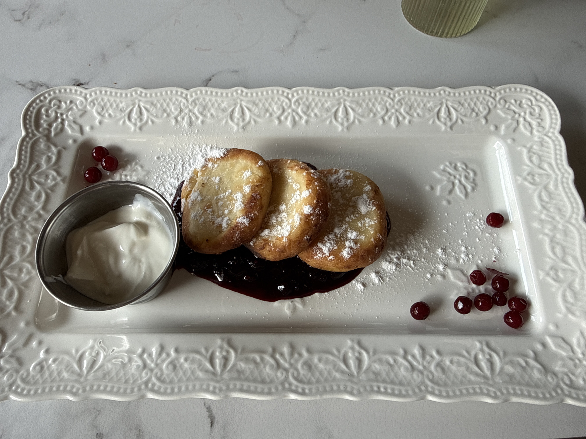 A plate of syrniki, a Russian cheese pancake, is served with a side of yogurt and berry jam. The syrniki are dusted with powdered sugar and garnished with fresh berries. The plate is white and ornate, with a floral pattern around the edges. The syrniki are stacked three high, and the jam is a dark, rich color. The yogurt is thick and creamy. The scene is lit from above, creating a soft, inviting glow. The location in the city of Ust'-Muny, Russia is not specified.