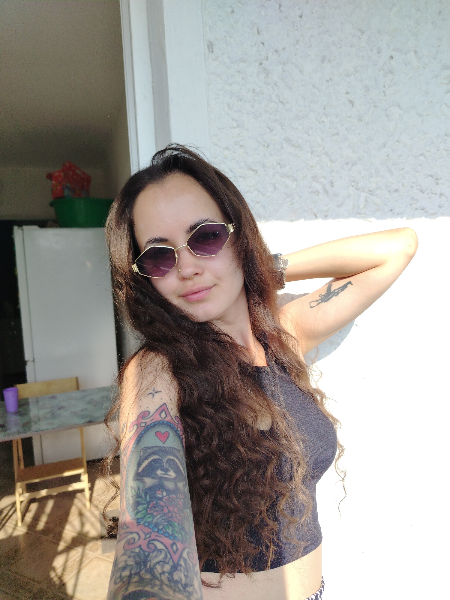 A young woman with long, dark, curly hair and sunglasses is taking a selfie outdoors. She is wearing a dark tank top and has tattoos on both arms. She is leaning against a white, textured wall, with her right arm behind her head. To her left, a table with chairs and a refrigerator can be seen in the background. The scene appears to be in a residential area, possibly a backyard or patio, with sunlight casting shadows on the ground. The woman's facial expression is neutral, and she is looking directly at the camera. The image captures a casual, candid moment.