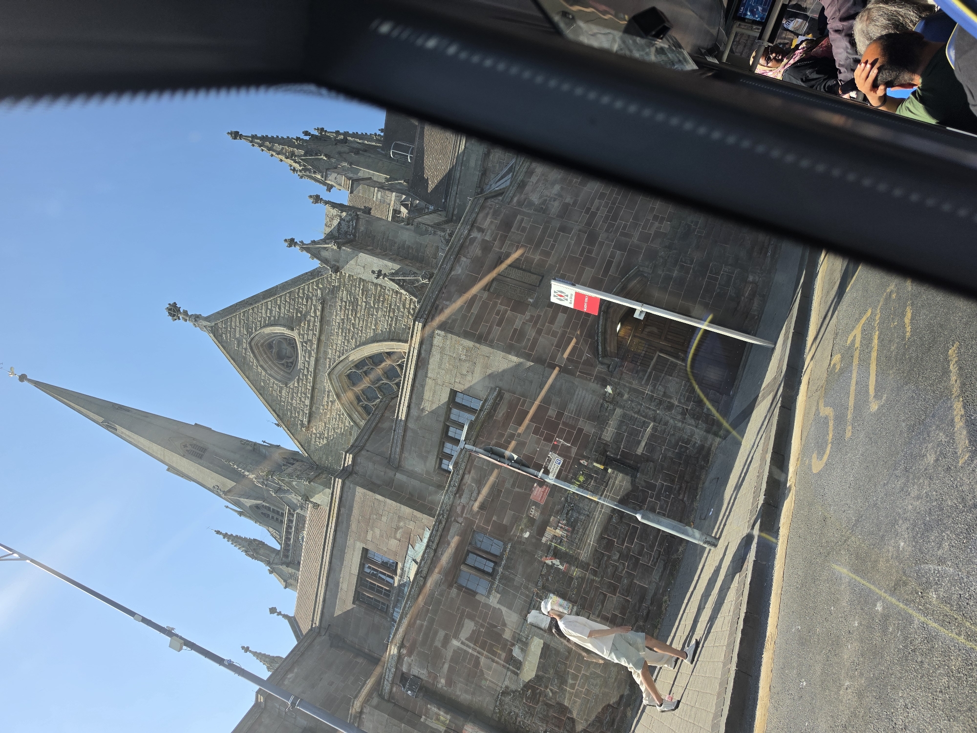 The image shows a view from inside a vehicle looking out at a large stone building with a tall spire, characteristic of a cathedral or large church. The building is made of dark, weathered stone blocks and features Gothic architectural elements such as pointed arches, decorative stonework, and multiple spires. The sun is bright, casting shadows and creating lens flare on the vehicle's window.
On the sidewalk in front of the building, a person wearing a light-colored t-shirt, light-colored shorts, and a white cap is walking. They are wearing white sneakers with pink accents. There is also a lamppost with signage on the sidewalk.
Through the window of the vehicle, several passengers are visible. One woman in the foreground is wearing a patterned headscarf and glasses. Another passenger, a man, is seen from behind, touching the back of his head. The interior of the vehicle appears to be a bus or a similar public transport, indicated by the dark frame and the presence of multiple passengers.
The road in front of the sidewalk has yellow markings, including a number '35' painted on the tarmac. The building's architecture strongly suggests it is St. Philip's Cathedral, located in the city center of Birmingham, United Kingdom.