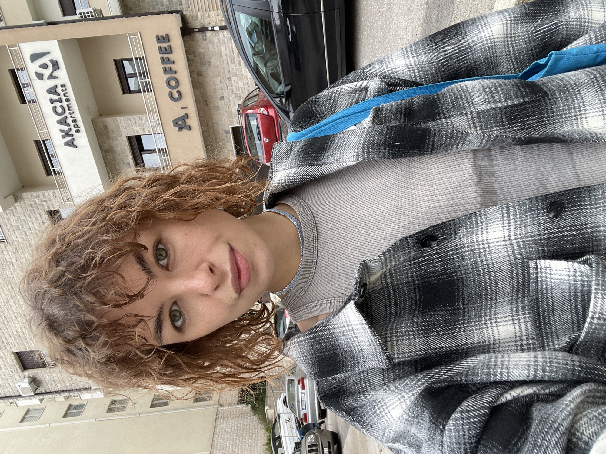 A young, fair-skinned woman with light brown, curly hair and green eyes looks directly at the camera. She is wearing a gray ribbed tank top and a black and white plaid overshirt with a blue strap underneath. Behind her is a building labeled "AKACIA apartments" and "A. COFFEE". There are several cars parked along the street. The location is Boreti, Montenegro, evidenced by the license plates on the parked vehicles.