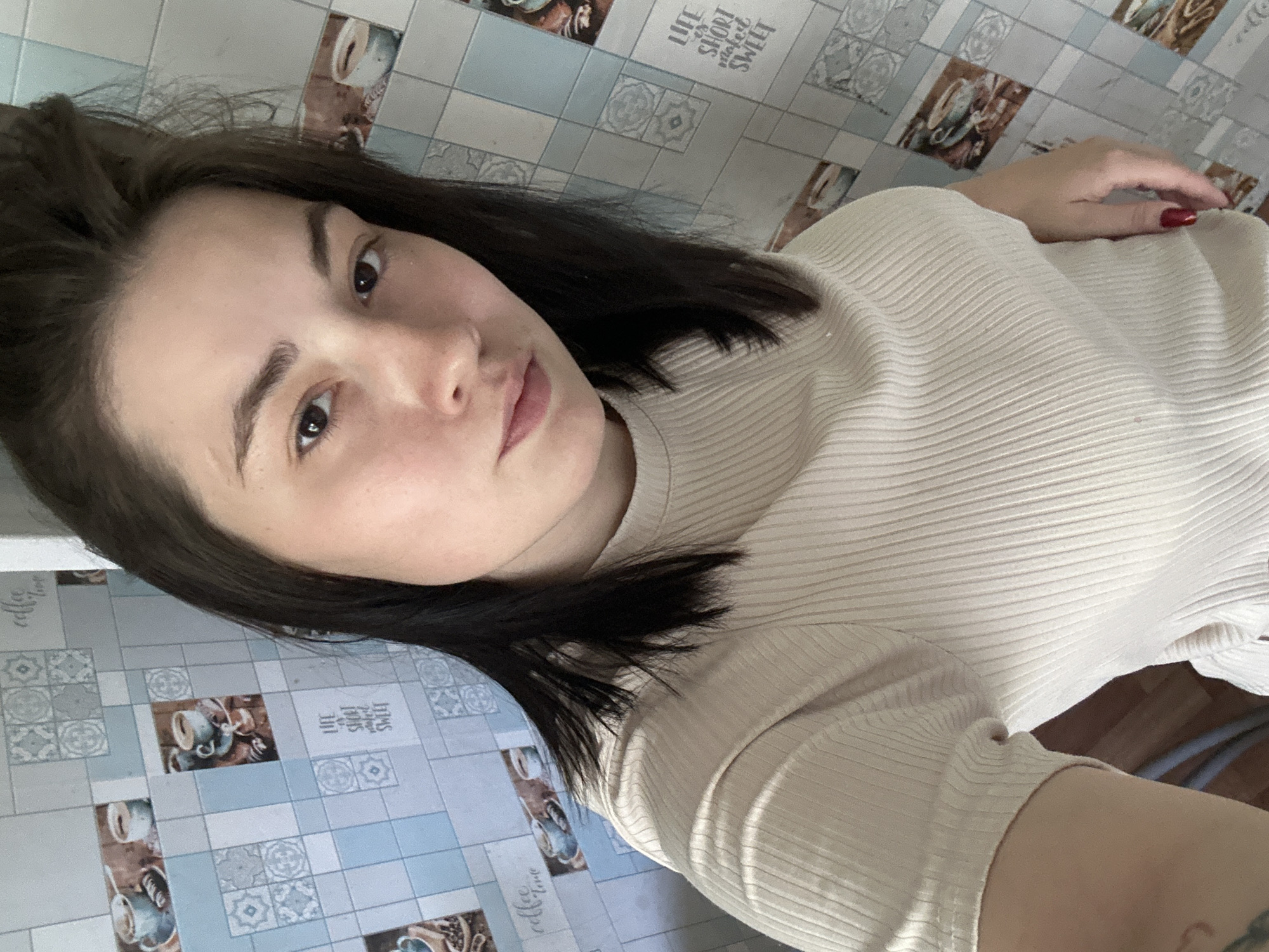 A young woman with dark hair is taking a selfie in what appears to be a kitchen or bathroom. She is wearing a beige ribbed t-shirt. The wall behind her is tiled with a pattern that includes images of coffee cups and text, some of which reads "coffee time" and "Life is short, make it sweet." The overall lighting is bright. The location is the city of Chaikovsky, Russia.