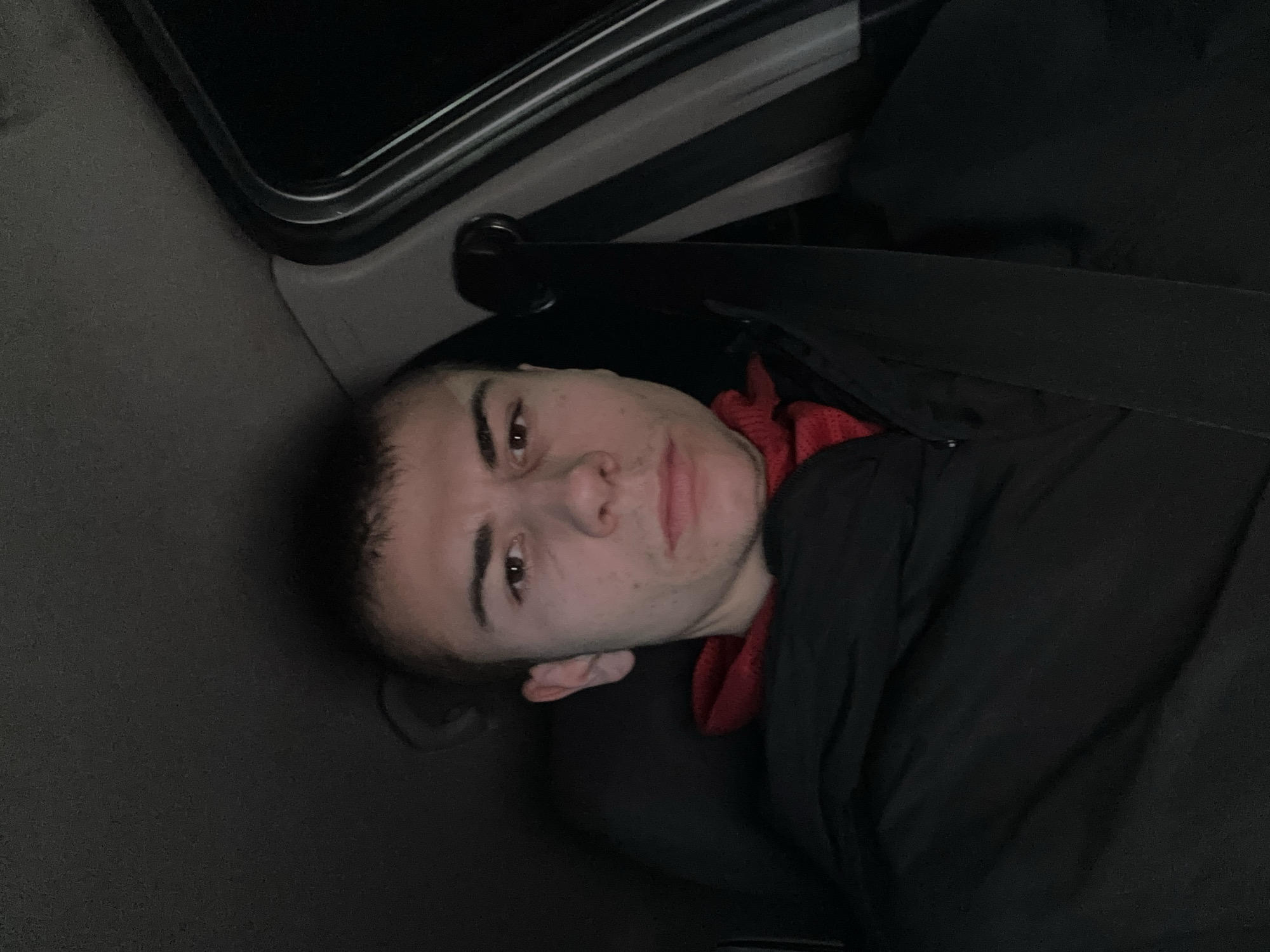 A young man is lying down in the back of a car. He is wearing a red garment under a black jacket, and his seatbelt is fastened. The location is likely within a vehicle in Plovdiv, Bulgaria.