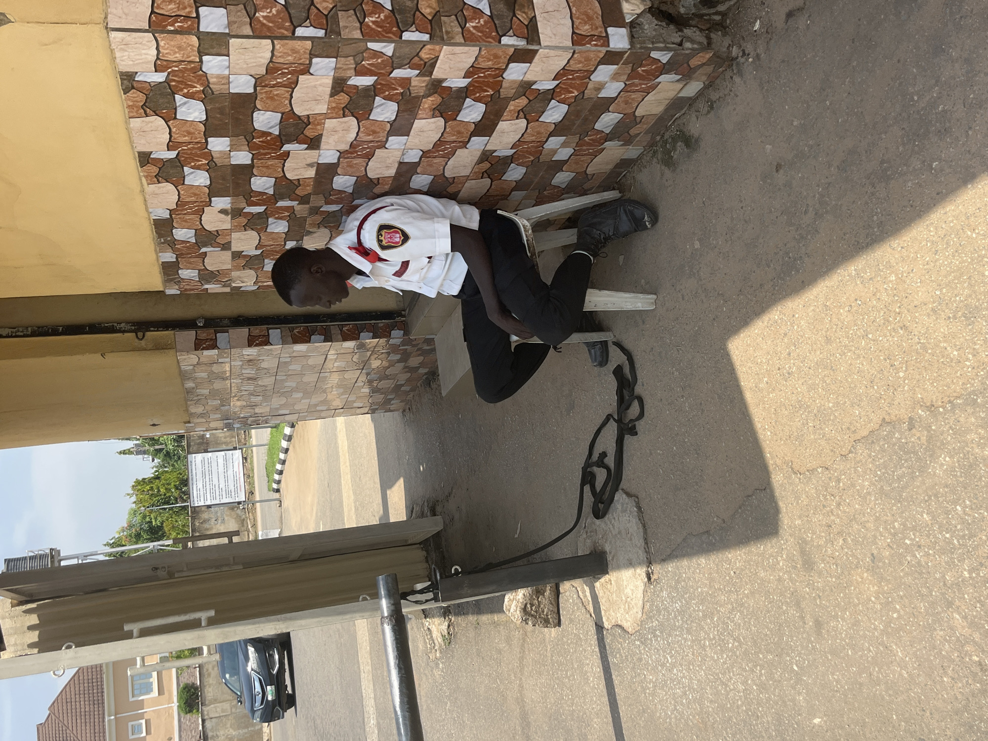 A young Black male in a security uniform, consisting of a white shirt with a red and gold emblem on the left breast and black trousers, is seated on a portable silver chair. He is leaning forward with his head bowed, appearing to be resting or asleep. His black boots are visible. A black strap or rope is coiled on the ground next to the chair.
The scene appears to be outdoors, in an area with a tiled wall and a paved ground surface. The tiles on the wall have a mosaic pattern of brown, white, and beige squares. Sunlight casts a shadow across the ground, suggesting it is daytime, likely late morning or early afternoon. In the background, there are glimpses of a building, some greenery, and a signpost with text. A dark-colored car is partially visible in the distance. The overall setting suggests an urban or suburban environment. There are no overt emotional expressions visible due to the subject's posture. The text on the signpost is not legible in detail.