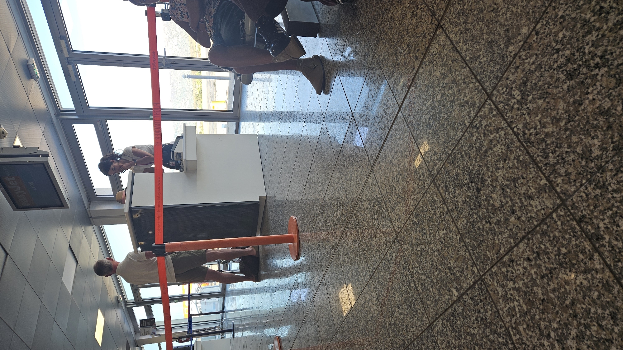 The image depicts an airport check-in or gate area. In the foreground, a man stands with his back to the camera, wearing a white polo shirt and khaki shorts, with a dark bag at his feet. To the right, a woman in a white polo shirt and dark skirt is standing behind a counter, appearing to be checking something on a mobile device. A straw hat rests on the counter. Red stanchion belts divide the waiting area. In the background, glass doors offer a view of an outdoor area with distant hills. A digital display screen above the counter shows "easyJet" and "LYON." The floor is tiled with a speckled granite pattern, reflecting the ambient light. A person is partially visible sitting to the far right, wearing a patterned shirt and shorts, with their legs crossed.