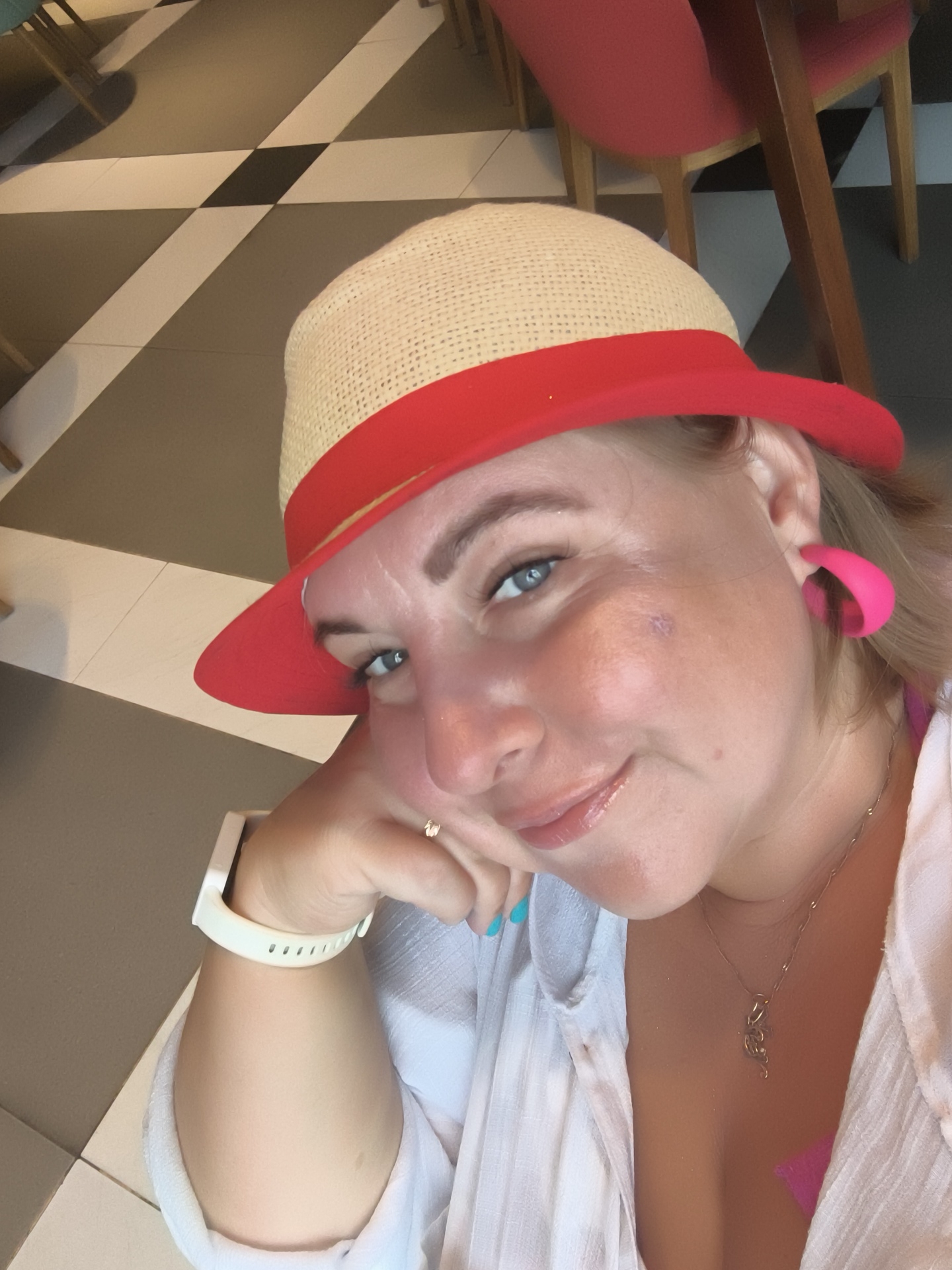 A woman wearing a straw fedora with a red band and brim poses for a selfie in a dimly lit room with a checkered floor and red chairs. She has bright blue eyes, is wearing a white shirt, and a pink earring. A white watch is on her left wrist, and she is wearing a necklace. The scene is likely indoors, possibly in a cafe or restaurant, given the furniture and flooring. The specific location is not discernible from the image alone; it is not possible to determine if the image was taken in Phú Quốc, Vietnam.