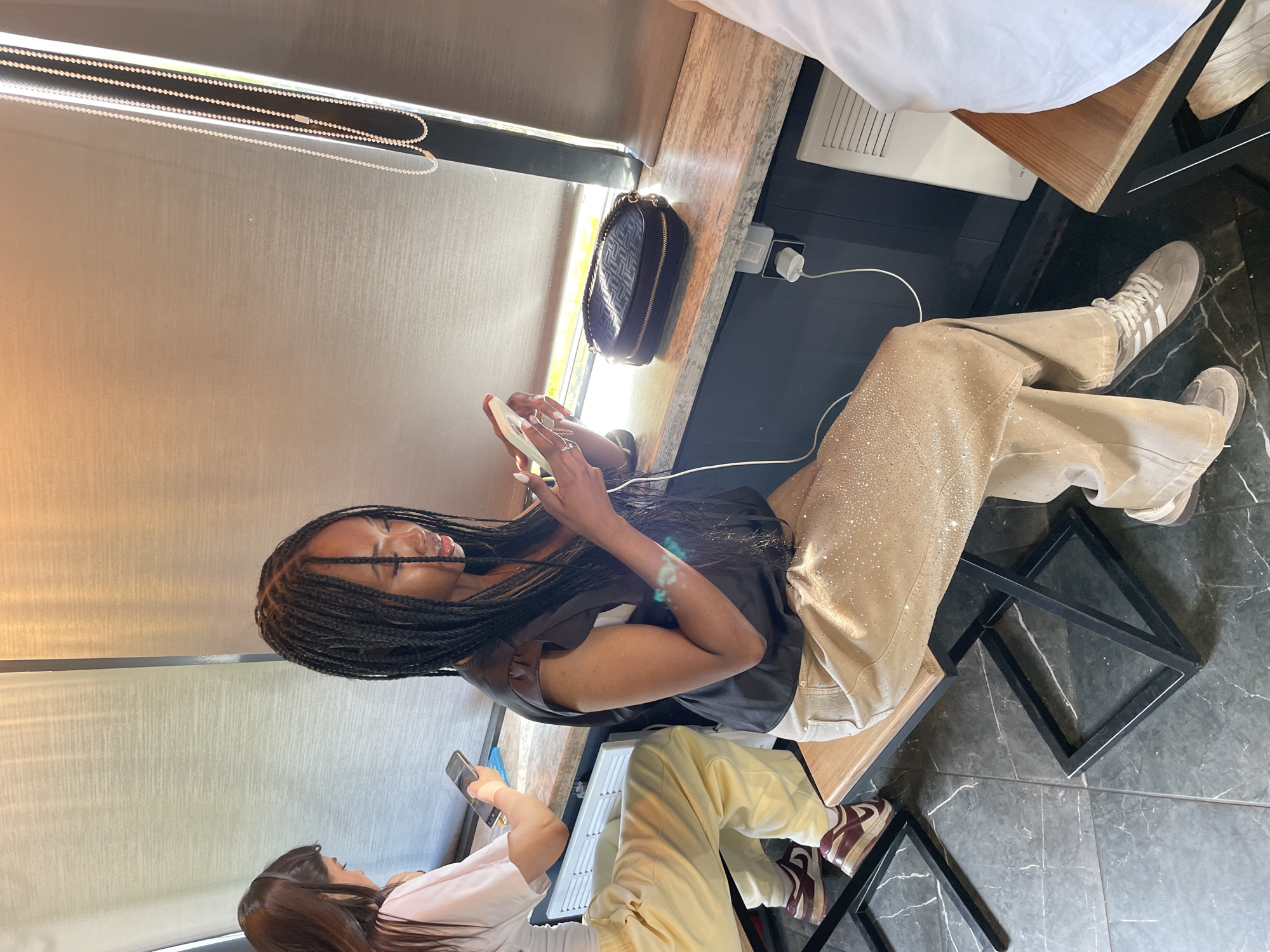Two young women are seated at a cafe. The woman in the foreground has dark skin and braids, wearing a black top and tan pants adorned with sparkles. She is looking at her phone. To her left, another woman with long brown hair, wearing a white t-shirt and yellow pants, is also looking at her phone. They are seated at a counter with a window behind them. The environment appears to be a cafe or a similar establishment.