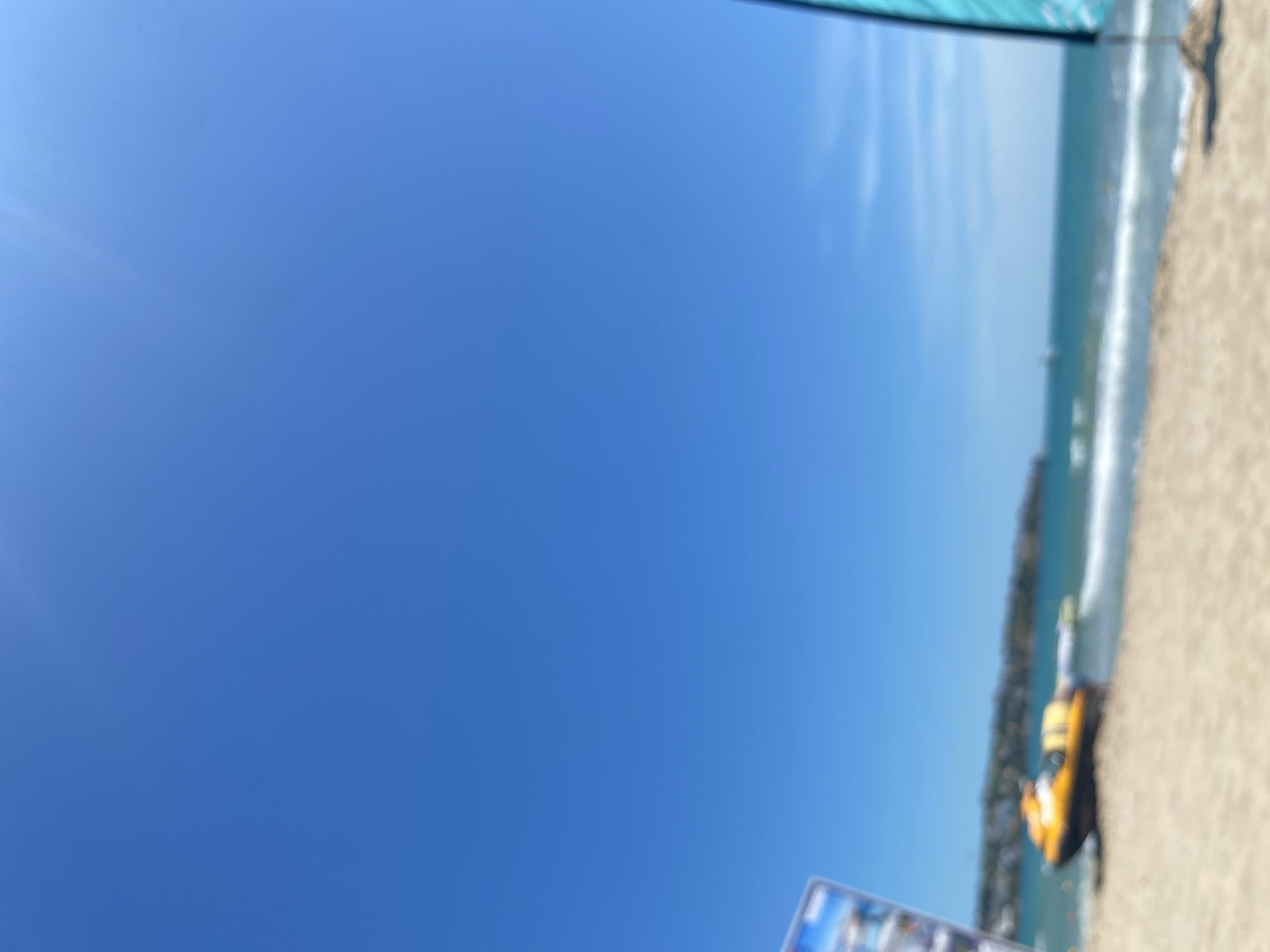 A blurred image of a beach scene on a sunny day shows a bright blue sky above a sandy beach and the ocean. In the foreground on the sand is a yellow jet ski. In the distance, land with buildings is visible along the coastline, and several small boats are in the water. A tall teal flag on a stand is positioned on the right side of the image.