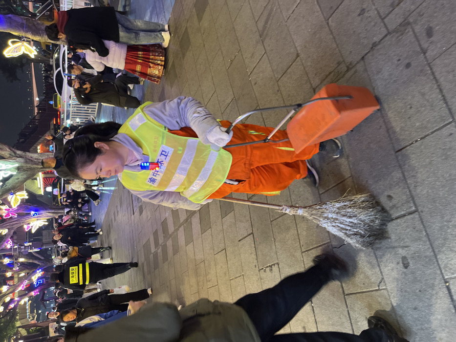 An outdoor, eye-level forensic image taken at night in Yuzhong District, China, shows a female sanitation worker as the primary subject on a paved pedestrian area. The woman, positioned slightly right of center in the foreground, wears a light grey long-sleeved top, bright orange overalls, and a neon yellow reflective vest with white horizontal stripes. The vest features Chinese characters "渝中环卫" (Yuzhong Sanitation) printed in blue. A small, multi-colored flashing device is clipped to her vest. She wears white gloves and holds a worn, dirty straw broom with a wooden handle in her right hand. In her left hand, she pulls an orange, rectangular refuse bin on wheels with a silver metal handle.
The immediate ground is a light grey paved surface composed of rectangular tiles. In the mid-ground and background, numerous people are present, indicating a busy public space. To the woman's left, a blurred individual's legs and torso are visible, walking past. Further back, a person wearing a black jacket with yellow reflective stripes and Chinese characters resembling "应急保安" (Emergency Security) walks away from the camera. To the woman's right in the mid-ground, a photographer in a dark jacket is kneeling, holding a camera, pointing towards two individuals posing: one person in a light-colored top and a vibrant red and gold patterned skirt, embraced by another person in a dark jacket and jeans.
The background is illuminated by decorative string lights on trees, featuring prominent glowing blue and purple butterfly shapes, as well as general city lighting. Faintly visible beyond a metal fence in the far background are vehicle lights, possibly from an elevated roadway or bridge, and other city structures. The overall scene suggests an urban environment, likely a market or tourist area, at night.