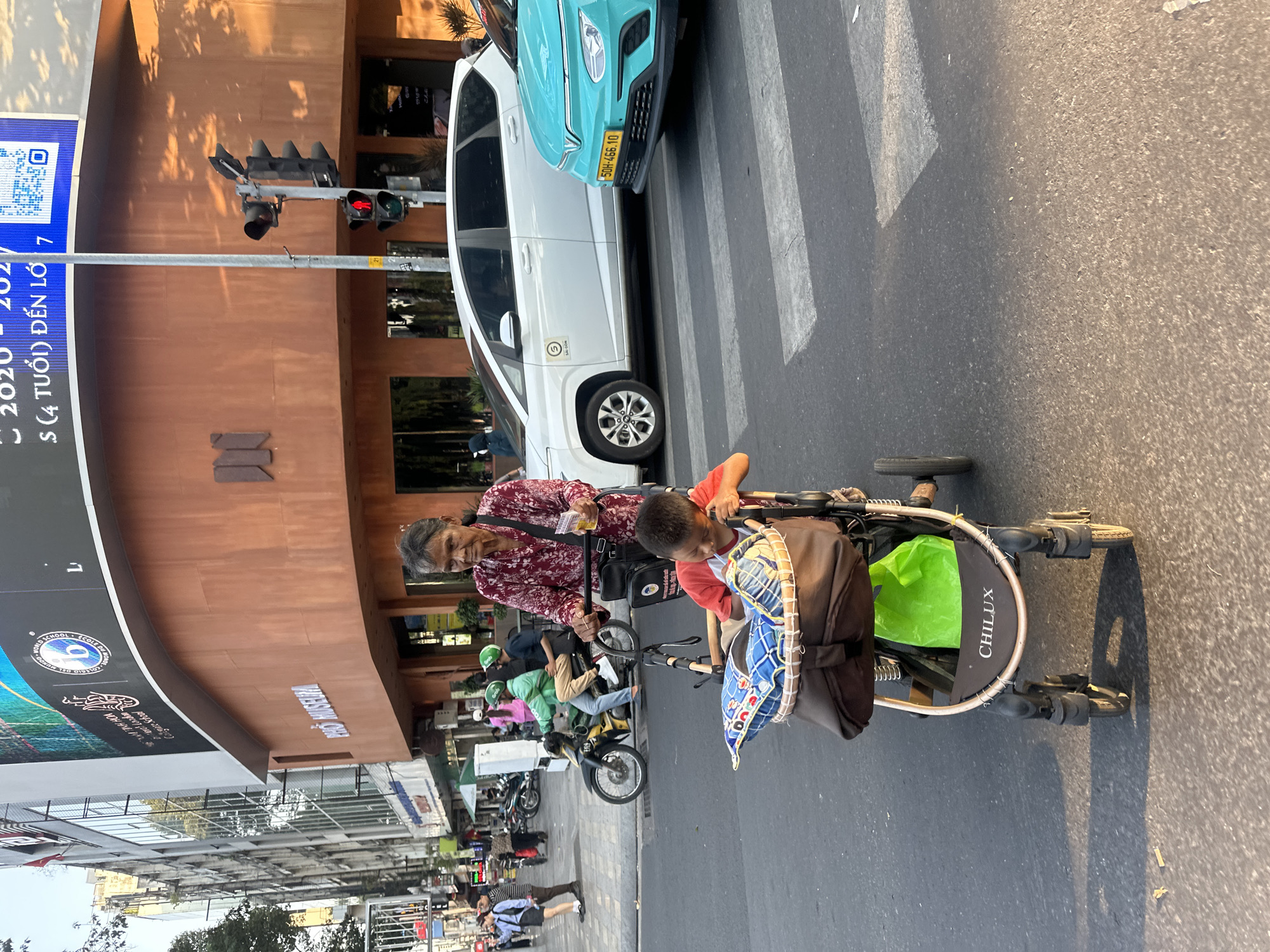 An elderly woman, of apparent East Asian ethnicity, with dark hair pulled back, is depicted pushing a stroller across a street on a marked zebra crossing. She wears a floral-patterned, long-sleeved shirt in red and white tones, and carries a black shoulder bag with a yellow item in her right hand. Seated in the dark-framed "CHILUX" brand stroller is a young boy, also of apparent East Asian ethnicity, dressed in a red short-sleeved shirt, leaning forward and looking down. A blue and white patterned pillow is positioned behind him, and a bright green plastic bag rests in the stroller's under-basket.
The scene is set in an urban environment, specifically a street intersection in what is specified as the city of Thủ Đức, Vietnam. In the background, a large building with a reddish-brown facade prominently displays "PHỞ MUSEUM" in white lettering. Above this building, a large digital billboard is visible, featuring text in Vietnamese and English, including "WORLD SCHOOL." To the left of the museum building, an older, multi-story structure with numerous windows is visible.
Vehicles are stopped on the asphalt road, consistent with a red traffic light visible on a pole. These include a white multi-purpose vehicle (MPV) with a "SÀI GÒN" sticker, and a light blue/teal electric VinFast VF e34 sedan with Vietnamese license plate "50H-466.10." Several motorcycles, some with riders wearing green helmets, indicating ride-sharing services, are also present. Pedestrians are visible on the sidewalk to the left, and shadows suggest late afternoon or early morning sunlight.