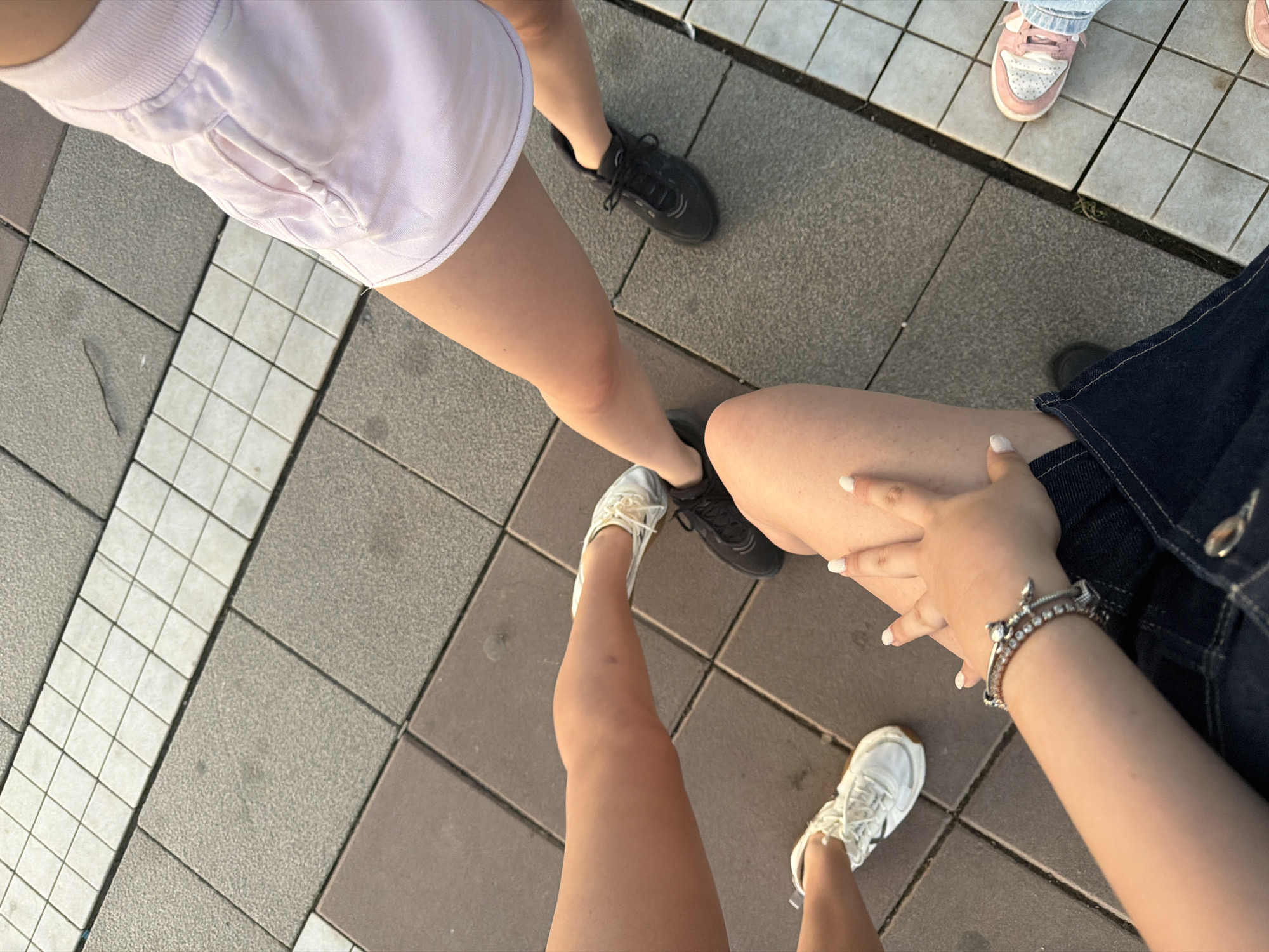 A high-angle forensic view captures the lower extremities of at least five individuals standing and sitting on a paved outdoor surface in Blagoevgrad, Bulgaria. The ground is composed of various square paving stones: large textured grey tiles, smoother dark brown tiles, and a distinct central band of smaller white square tiles.
From the top right, a person is visible wearing light pink athletic shorts, exposing bare legs, and black athletic shoes. Adjacent, to the left, is another bare leg with a small bruise noticeable on the shin, accompanied by white sneakers with metallic accents. Further left, a third set of bare legs and white sneakers with a distinctive brown sole are present. In the foreground, a person is seated or kneeling, identifiable by a bare leg, and a hand with white-painted fingernails resting on their thigh. This individual wears a silver-toned bracelet with decorative elements and dark denim clothing. Partially visible at the bottom right are light blue jeans and pink and white sneakers belonging to a fifth person. The scene is illuminated by natural daylight, casting soft shadows.