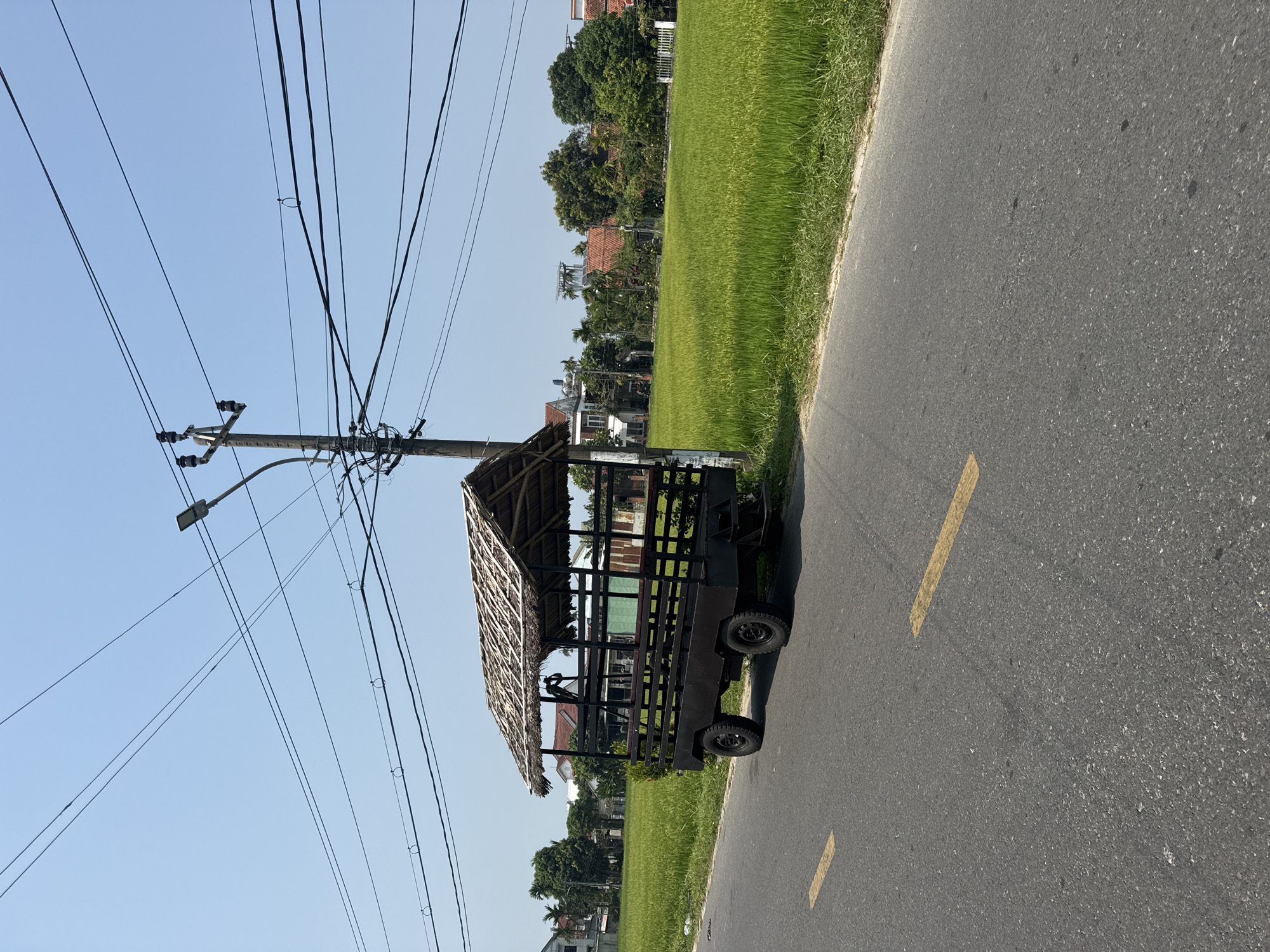 The image shows a utility cart with a thatched roof parked on the side of a paved road. A wooden utility pole with power lines runs along the road. Next to the road is a field of green grass. A few houses and trees can be seen in the background. The sky is clear and blue. The cart is situated in Đà Nẵng, Vietnam.