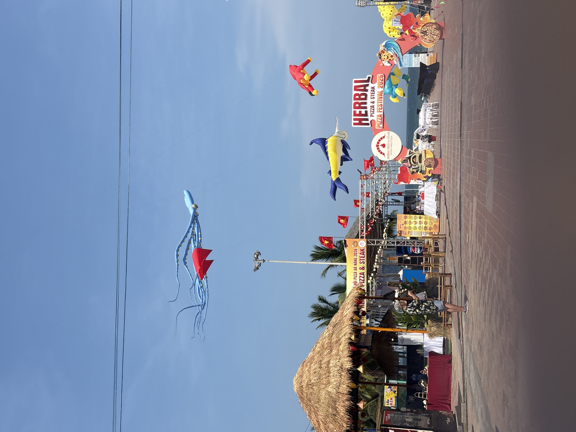 A vibrant outdoor festival is captured from a slightly elevated perspective, showcasing a clear blue sky adorned with several large, colorful kites. The scene depicts what appears to be the "HERBAL PIZZA FESTIVAL 2026" or "HỘI PIZZA ĐÀ NẴNG 2026," as indicated by prominent banners.
In the sky, a light blue octopus-shaped kite with red triangular tips, a red humanoid-shaped kite, and a yellow and blue swordfish-like kite are flown, adding a playful and festive atmosphere. Below, a paved promenade stretches into the distance, lined with various festival setups. On the right side, large decorative arches and banners mark the entrance or a section of the festival. Several Vietnamese flags (red with a yellow star) are visible, hanging from structures.
Along the promenade, there are thatched-roof stalls or huts, suggesting food vendors or exhibition booths. One such hut is prominent in the foreground on the left. Palm trees are scattered throughout the background, hinting at a tropical or coastal setting, further supported by the "ĐÀ NẴNG" (Da Nang) mention on a sign. A single person is seen standing on the paved area, facing away, observing the festival. The day is bright and sunny, with no clouds in the sky.
Visible text on various signs and banners includes: "HERBAL PIZZA & STEAK," "PIZZA & STEAK," "WOOD FIRED PIZZA," "HERBAL PIZZA FESTIVAL 2026," and "HỘI PIZZA ĐÀ NẴNG 2026." A smaller sign also reads "GAP BIM." The overall setting suggests a lively daytime event, likely near the ocean in Da Nang, Vietnam.