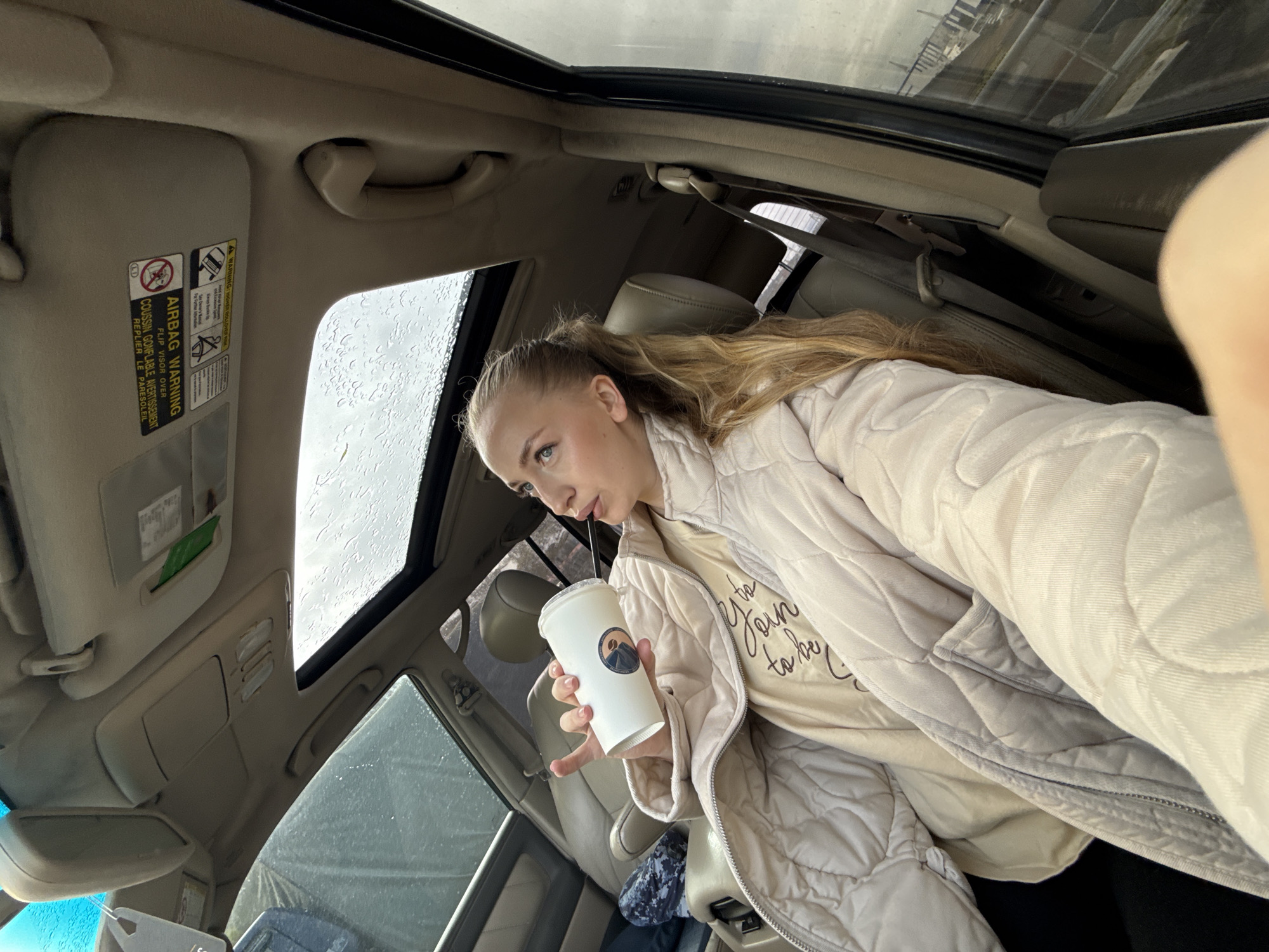 The image shows a woman in a car, possibly taking a selfie. She is wearing a light-colored quilted jacket over a beige t-shirt with the words "join to be you" printed on it. She has her blonde hair pulled back in a ponytail and is holding a white paper cup with a black straw. The car has a sunroof, visible with raindrops. The sun visor on the driver's side is down, displaying an airbag warning label.
Based on the limited information, I cannot determine the precise location in Petropavlovsk-Kamchatsky, Russia.