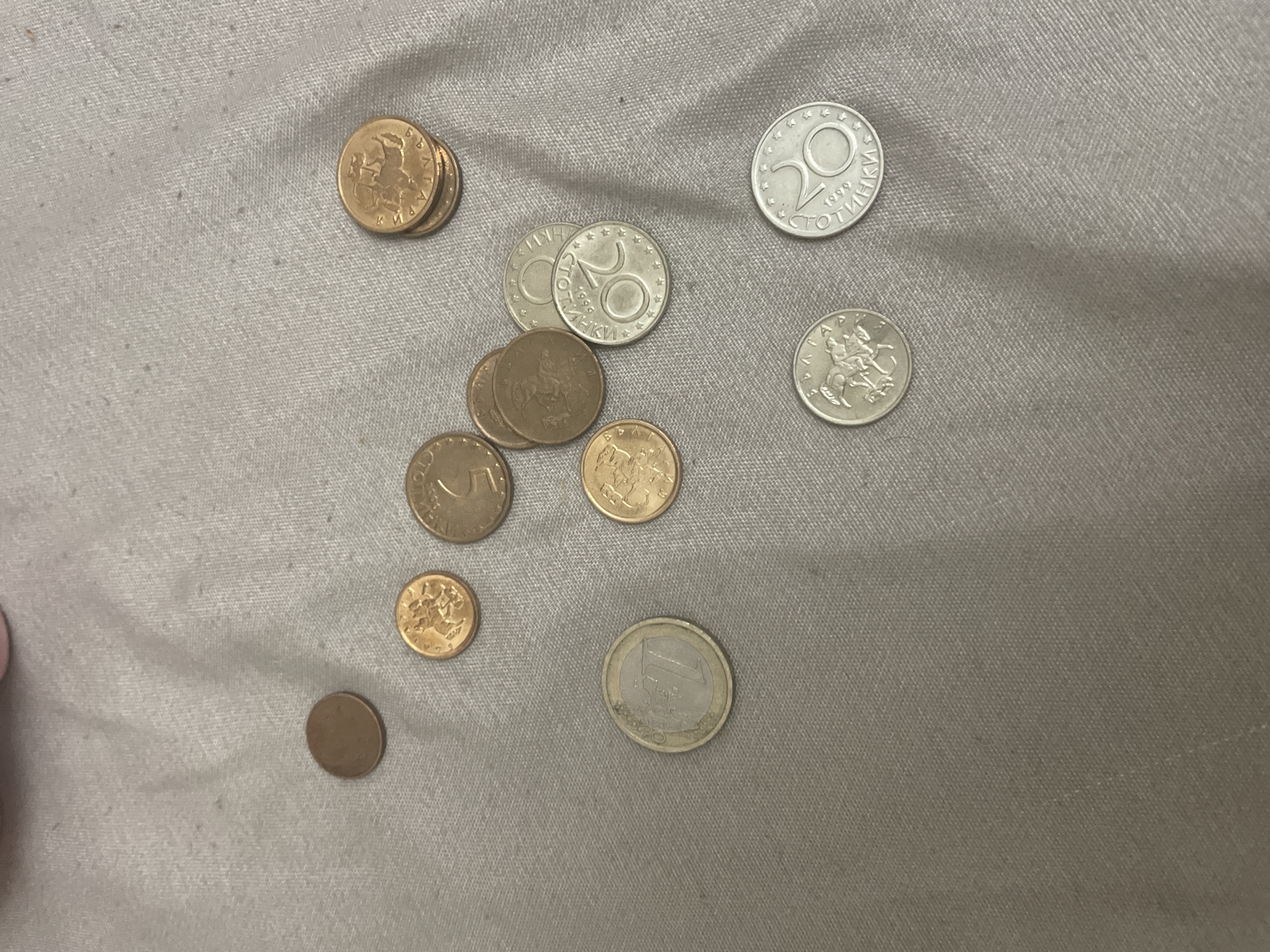 The scene is situated in Ihtiman, Bulgaria. The image depicts a collection of fourteen coins of various currencies and denominations, scattered across a light grey, textured fabric surface that exhibits minor wrinkles and a fine woven pattern. No individuals are present.
Identifiable coins include:
* One bi-metallic 1 Euro coin, dated 2002, located in the lower-left quadrant.
* A stack of three gold-colored Lithuanian Litas coins in the upper-right area, with the topmost coin clearly showing the Vytis (knight on horseback) and inscriptions "LIETUVOS LITAI 1999".
* Two gold-colored Bulgarian 50 Stotinki coins, one partially obscured below the Litas stack, the other showing a "5" and a lion design.
* Three Bulgarian 20 Stotinki coins, two silver-colored and one gold-colored, each dated 1999 with a lion design and "СТОТИНКИ" inscription.
* Two Bulgarian 10 Stotinki coins, one silver-colored and one gold-colored, both featuring a lion design.
Additionally, one small brown-colored coin is located in the upper-left, and two other small gold-colored coins are positioned in the upper-middle and upper-right areas; their specific denominations and currencies are not clearly discernible due to resolution. The fabric surface serves as the sole environment, suggesting an indoor setting.