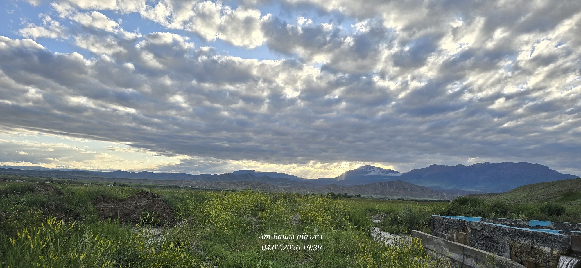 A landscape photograph captures a field of green grass and yellow wildflowers with a dirt pile in the foreground, leading to a body of water. In the distance, rolling hills and mountains are visible under a cloudy sky with rays of sunlight breaking through. A concrete structure with blue accents is present on the right side of the frame. Text overlay in a light color reads "At-Bashy айылы 04.07.2026 19:39". The scene appears to be an outdoor environment in the village of At-Bashy, Kyrgyzstan.