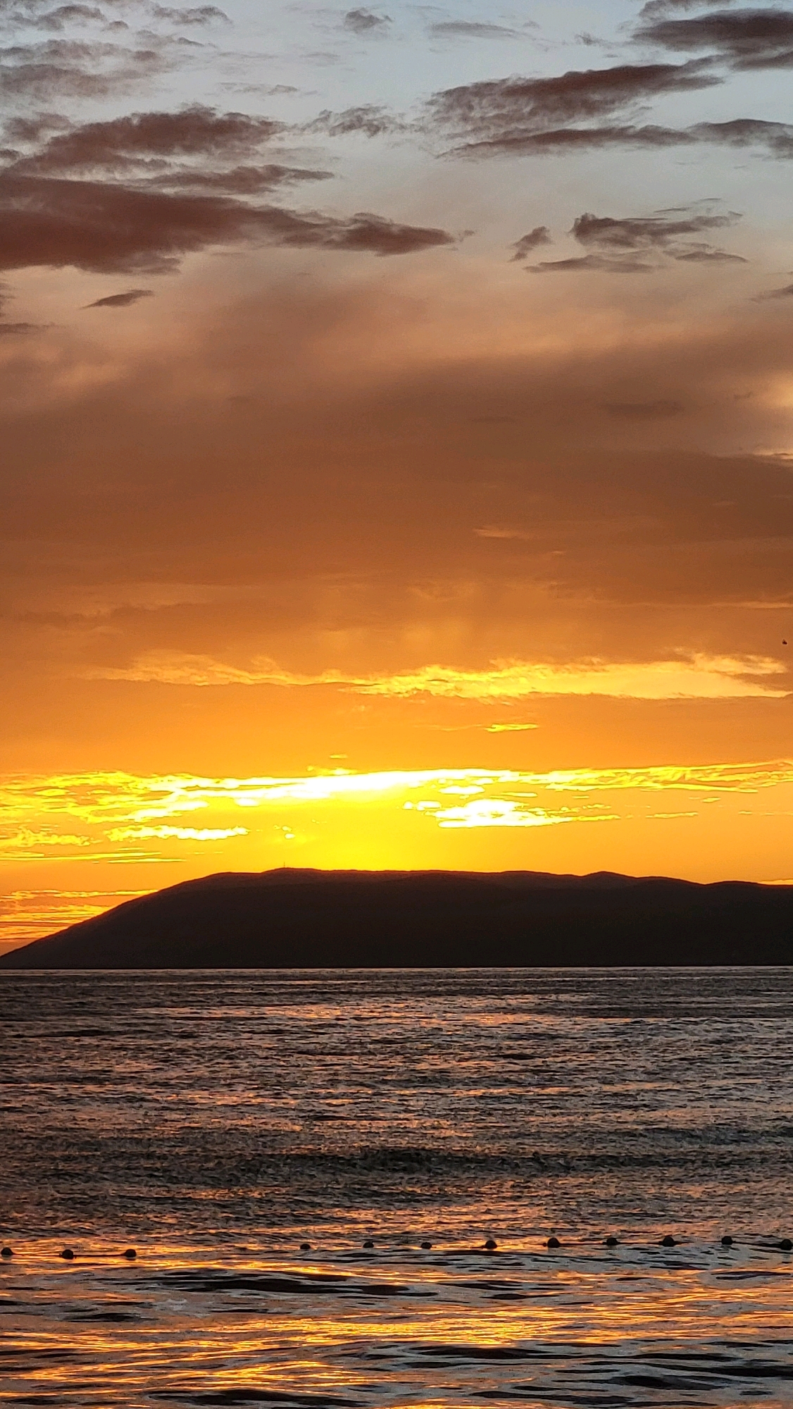 The image depicts a sunset over the sea, with a dark, silhouetted mountain range in the background. The sky is filled with clouds, displaying a gradient of colors from light blue at the top to bright orange and yellow near the horizon, indicating the setting sun. The sea reflects the warm hues of the sky, creating shimmering patterns on its surface. In the foreground, a line of buoys is visible on the water. The scene suggests a coastal location, possibly along the Makarska Riviera in Općina Tučepi, Croatia, known for its beautiful Adriatic coastline and mountainous backdrop. There are no people or visible actions in the image; it is a serene landscape shot.