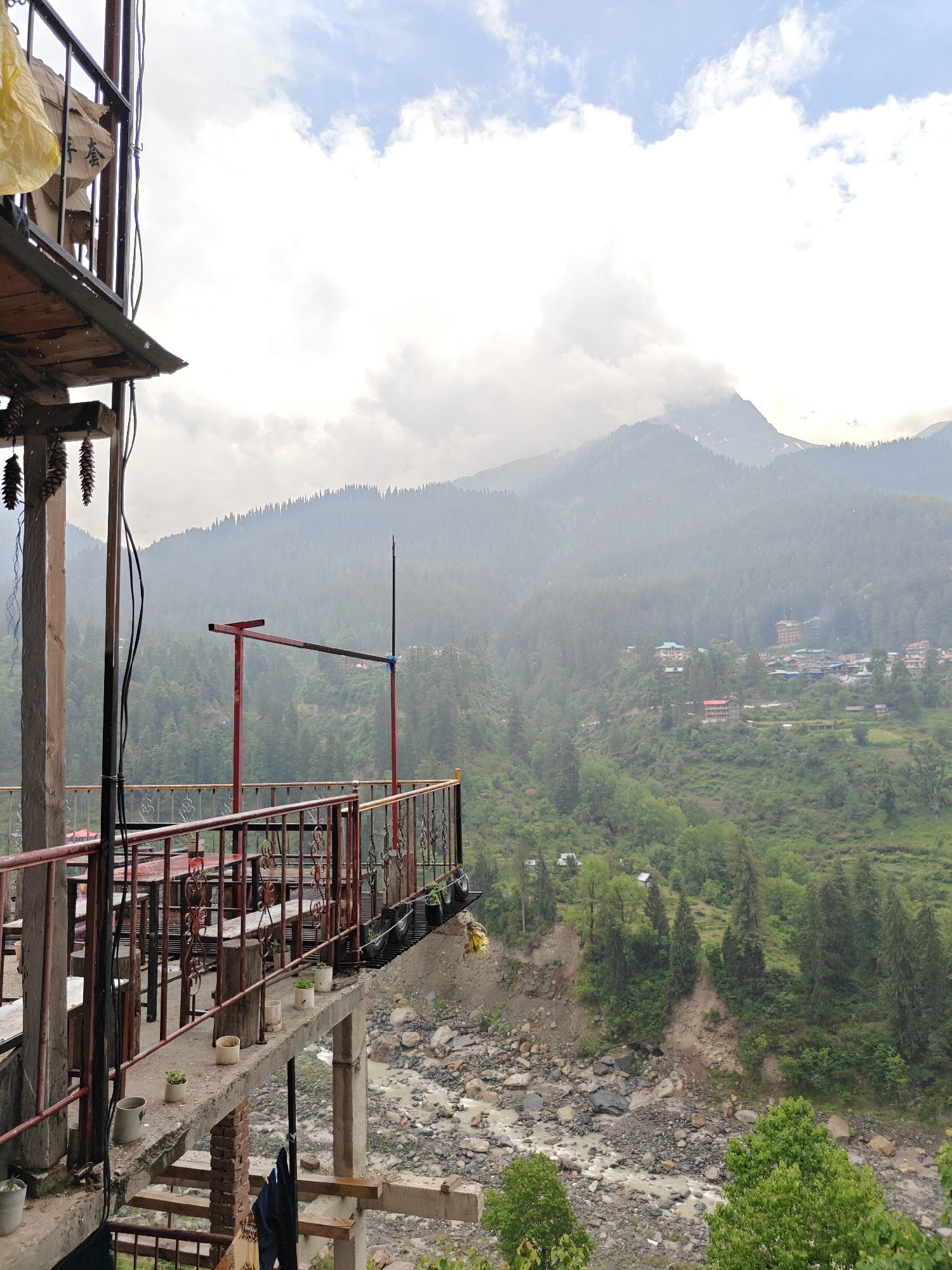 This photograph captures a panoramic view of a lush green mountain valley in Pulga, India, from the perspective of a multi-level outdoor terrace or balcony. In the immediate foreground, a rustic structure, likely part of a guesthouse or cafe, features wooden platforms, concrete supports, and a prominent reddish-brown metal railing with intricate scrollwork. Outdoor tables and benches are visible through the railing, set for patrons. Small potted plants line the balcony ledge, and decorative pinecones hang from an overhead wooden beam on the left. Electrical wires run down the side of the structure, and clothes are seen drying on a lower level. A yellow plastic bag and a brown sack, partially inscribed with Chinese characters (e.g., "营 套"), are suspended from the upper left.
Beyond the railing, the ground drops steeply to a rocky riverbed where murky water flows through. The opposite hillside is densely covered in coniferous forests, extending up majestic mountains. A small village with several buildings is nestled into this distant green slope. In the background, higher mountain peaks are partially shrouded in atmospheric haze and soft clouds, characteristic of the Himalayan region.
The sky is a mix of overcast grey and brighter white clouds, with patches of blue visible, suggesting a cloudy but well-lit daytime. The scene evokes a sense of tranquil natural beauty and remote charm. No people are visible, allowing the expansive landscape and the rustic viewpoint to dominate the frame.