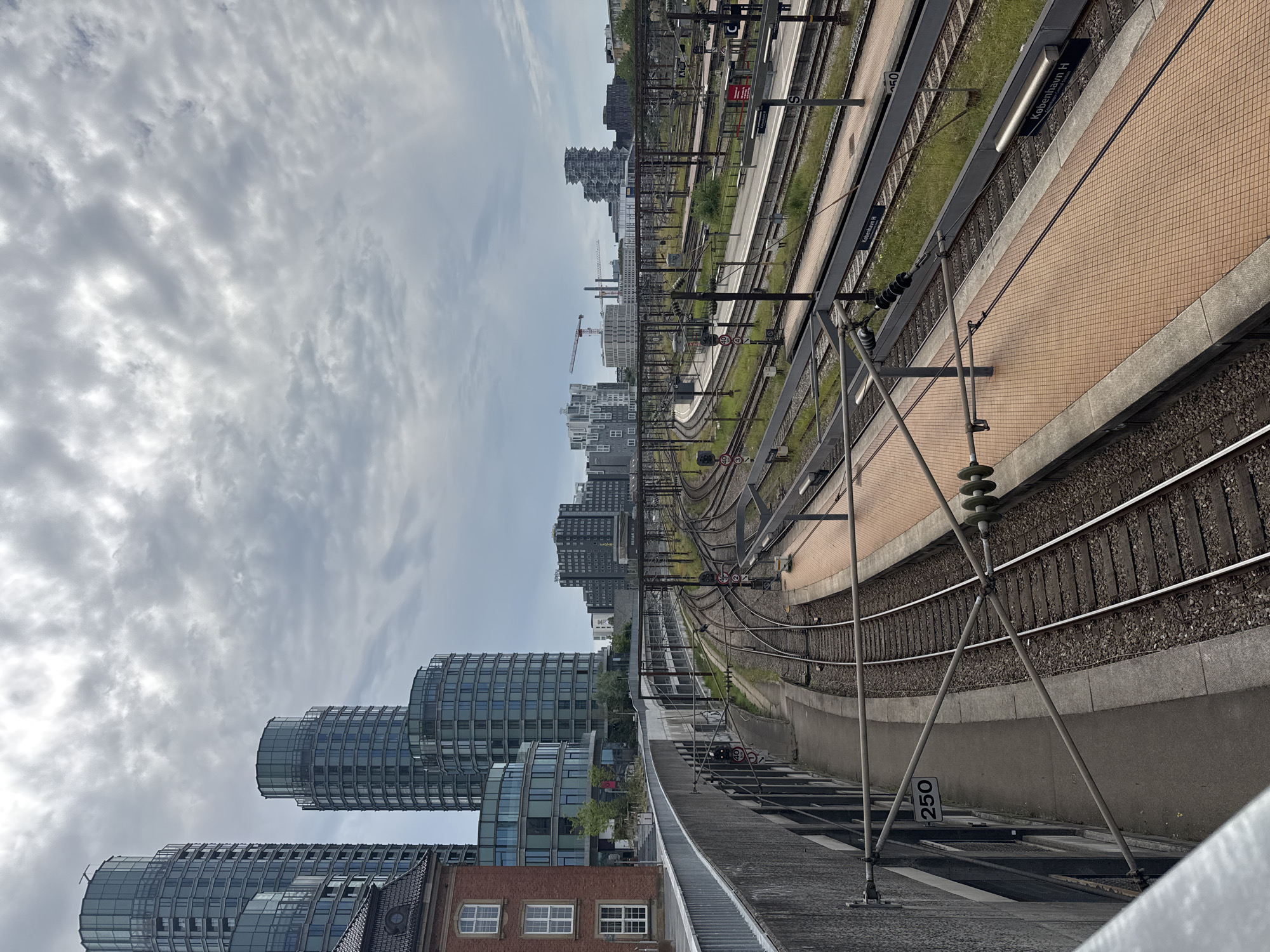 This image captures a view of the Kbenhavn H train station in Copenhagen, Denmark, looking out over multiple railway tracks and platforms. In the background, modern skyscrapers with rounded glass facades and angular designs dominate the city skyline. A red construction crane is visible in the middle distance. In the foreground, concrete structures, railings, and overhead power lines for the trains are prominent. The platforms are paved with orange tiles, and signs indicate "Kbenhavn H." The sky is overcast with cloudy, grey conditions. There are no people visible in the frame.