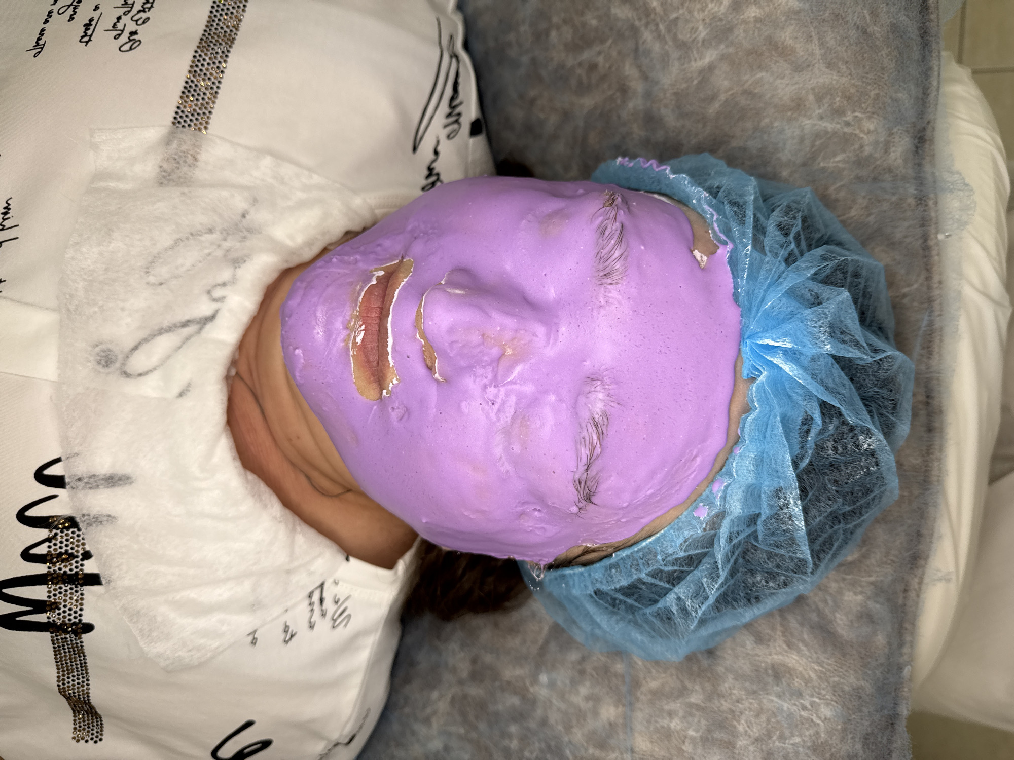 A top-down shot depicts a person lying down wearing a light purple facial mask. Some of the mask has peeled away near the mouth area, revealing the skin underneath. The person is wearing a white covering on their chest and neck, as well as a blue hair net on their head. The person appears to be on a gray surface. The location is Unknown, Unknown.