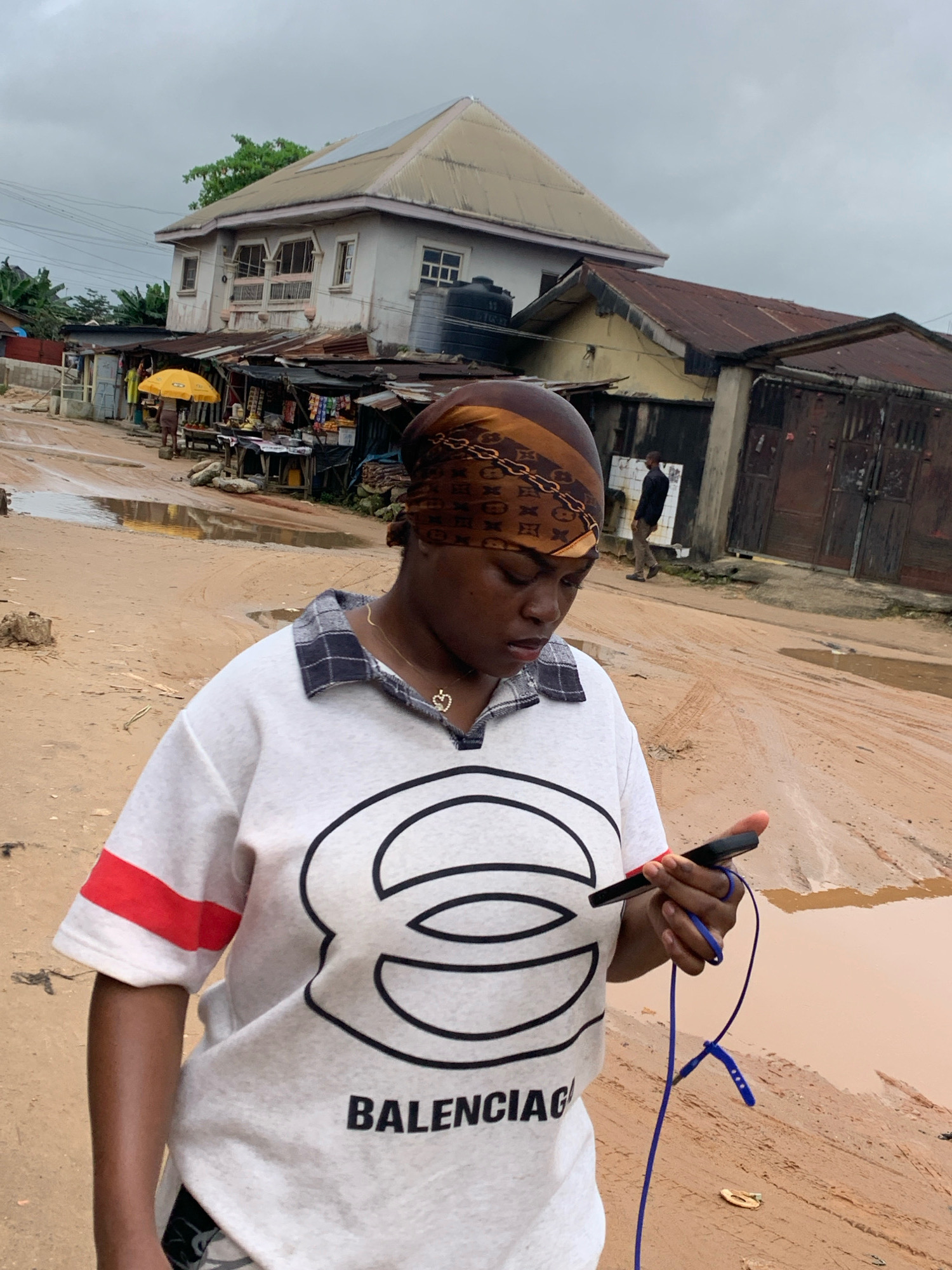 A young Black woman stands in the foreground of a muddy street, looking down at her smartphone. She is wearing a white t-shirt with a large black logo, a plaid collar, and a headscarf. In the background, several dilapidated buildings with corrugated metal roofs line the street. A person in a dark shirt and light pants walks away from the camera in the distance. The ground is covered in thick mud and puddles, suggesting recent rain. The sky is overcast. The scene appears to be a market or residential area in Umuode, Nigeria.