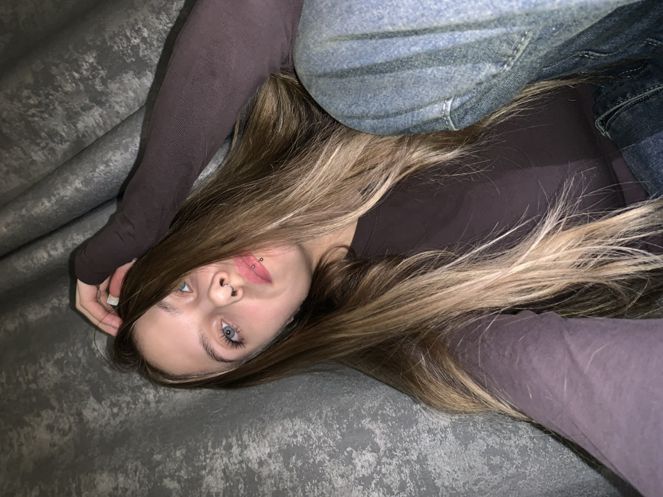 A close-up portrait features a young female positioned against a textured, mottled grey background, consistent with an indoor setting in the city of Фоки, Russia. The subject has long, multi-toned brown and blonde hair, styled with a center part. Her blue eyes are directed upwards and slightly to the viewer's right. She has a septum piercing in her nose and two labret piercings (snake bites) on her lower lip. She is wearing a dark brown long-sleeved top and blue denim jeans. Her left hand is raised, with fingers touching the hair on the left side of her head. The lighting is moderate, illuminating her face from the front.