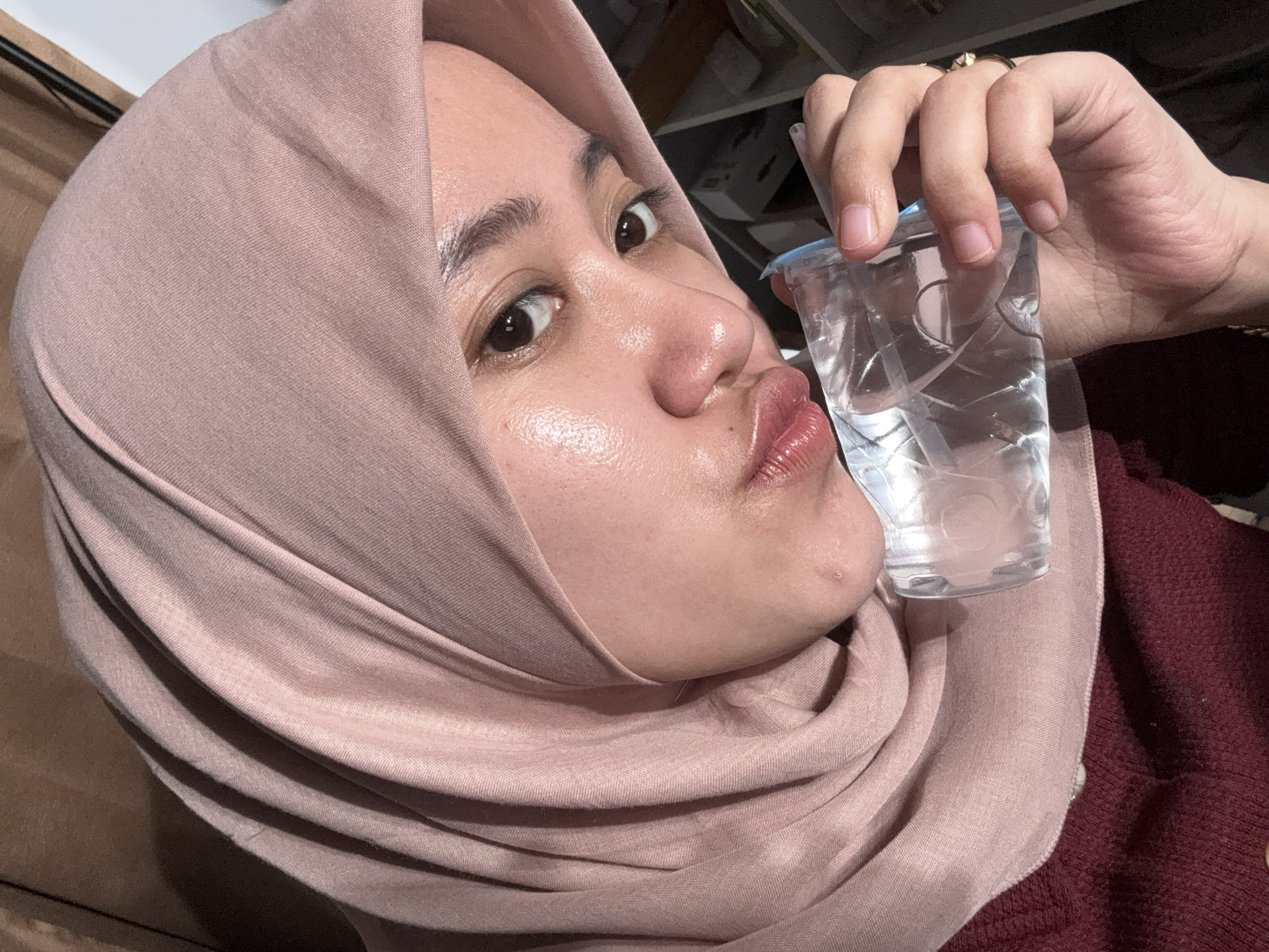 A woman wearing a pink hijab is holding a clear plastic cup filled with water. Her lips are pursed in a kissing or pouting expression. She has brown eyes, and the skin on her face appears slightly shiny. She is wearing a maroon-colored sweater. The background is blurry but suggests an indoor setting, possibly with shelving or storage units visible. The location is Gadingrejo, Indonesia.