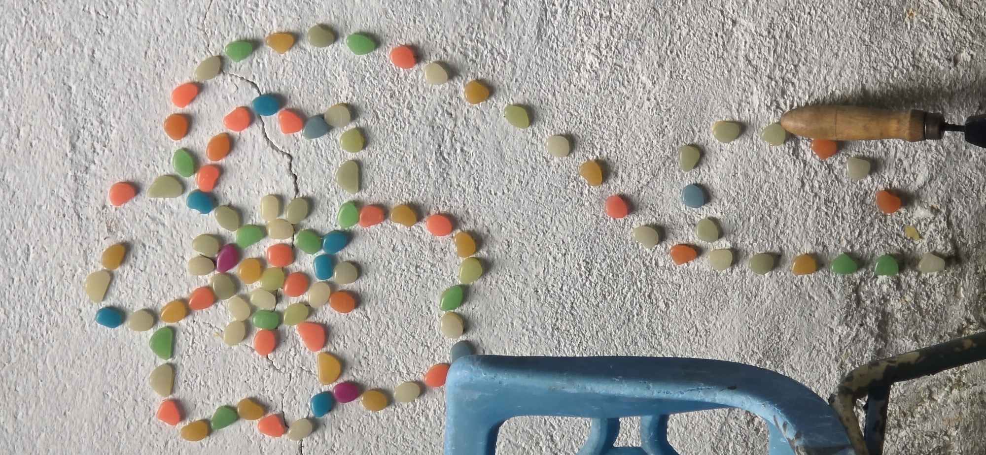 A white, textured wall surface in Partizanin, Bulgaria, is depicted, adorned with an arrangement of numerous small, smooth, colorful, pebble-shaped objects. These objects, exhibiting a variety of hues including light yellow, orange, pink, light green, blue, and purple, are meticulously placed to form distinct patterns: an abstract, stylized floral or star-like cluster in the upper-left quadrant and a larger, incomplete heart shape positioned in the center-right, from which a vertical trail of pebbles extends towards the bottom. The white wall displays visible cracks, particularly in the mid-right section. In the lower-left corner of the frame, a segment of a vibrant blue plastic chair or stool is present, showing part of its structure and a dark, possibly metallic, supporting frame below. In the lower-right, the light brown, wooden handle of a small hand tool, resembling a trowel, is visible. No individuals are present, but the deliberate arrangement of the pebbles indicates human interaction.
